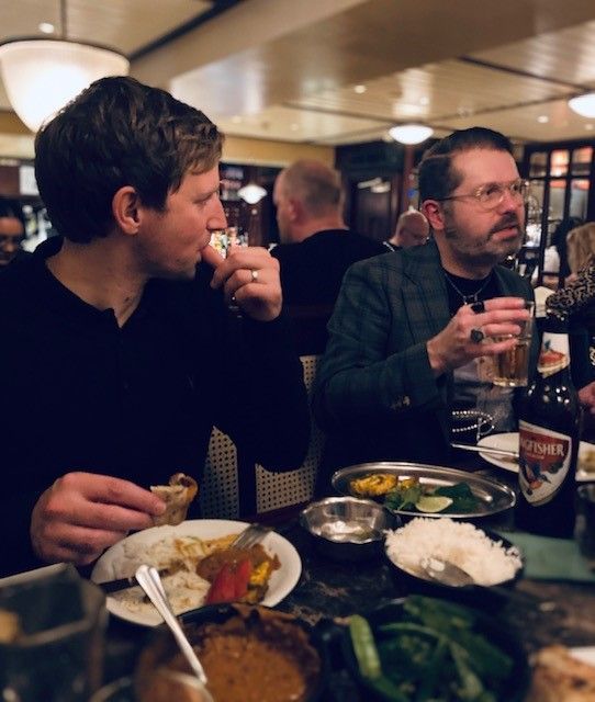 Two men eating Indian food in a restaurant. One man is drinking beer, the other eating with his hands.