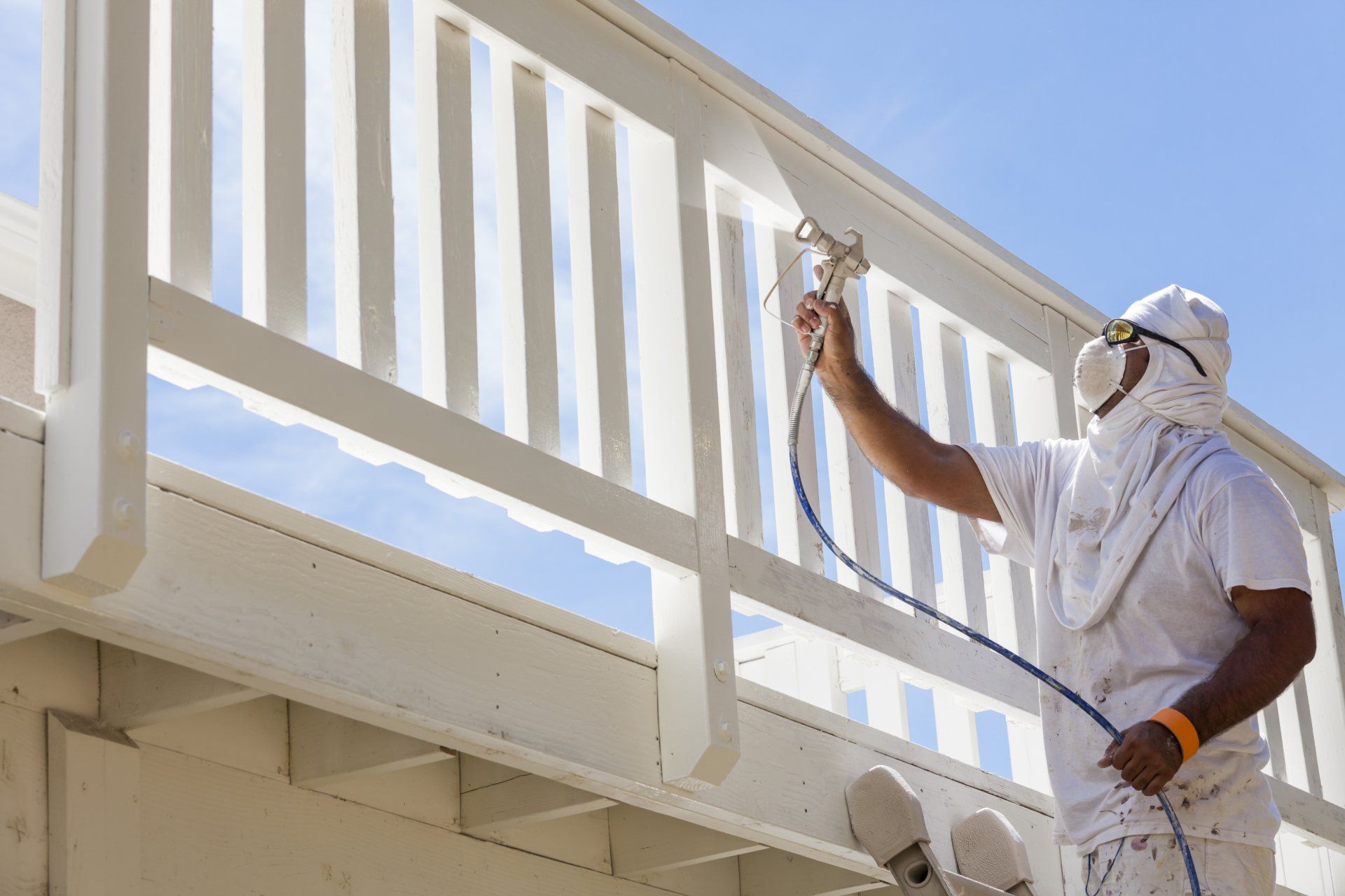 Person in protective gear sprays white paint on a wooden railing outdoors.