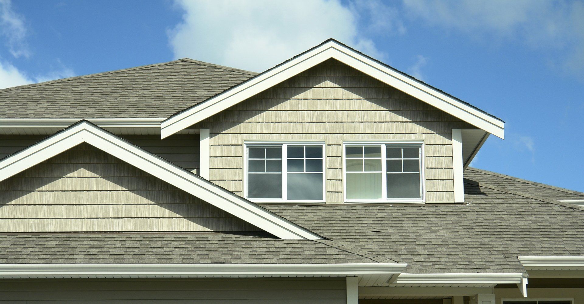 Tan shingled house roof with dormer windows against a blue sky.