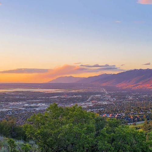 Cityscape at sunset with mountains, a lake, and trees in the foreground.