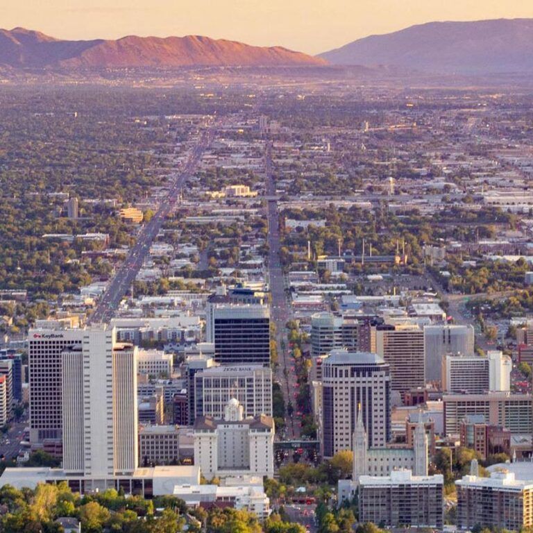 View of Salt Lake City, Utah, with downtown buildings, streets, and mountains in the background under a warm, golden sky.
