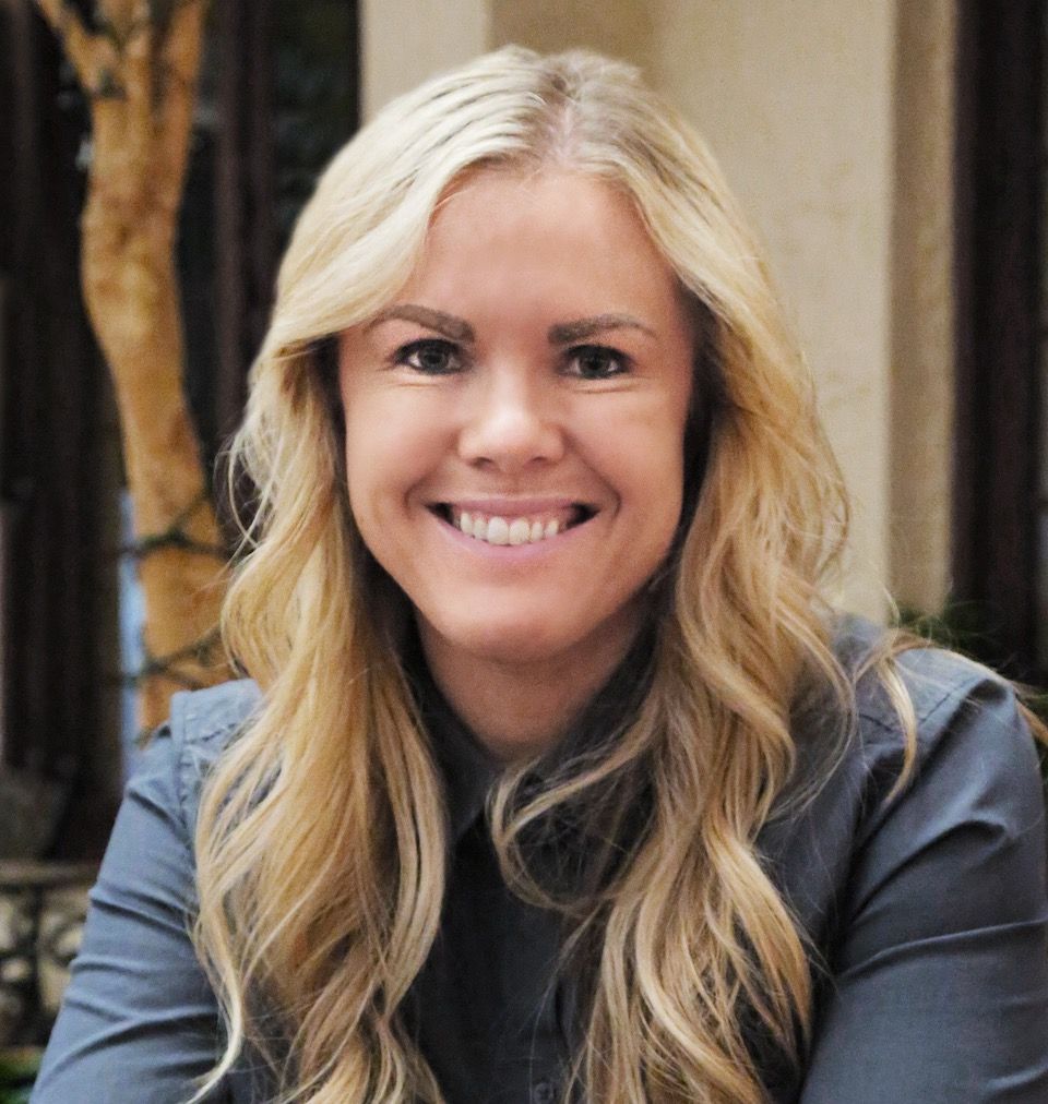 Blonde woman smiling, wearing a gray collared shirt, outdoors with blurred background.