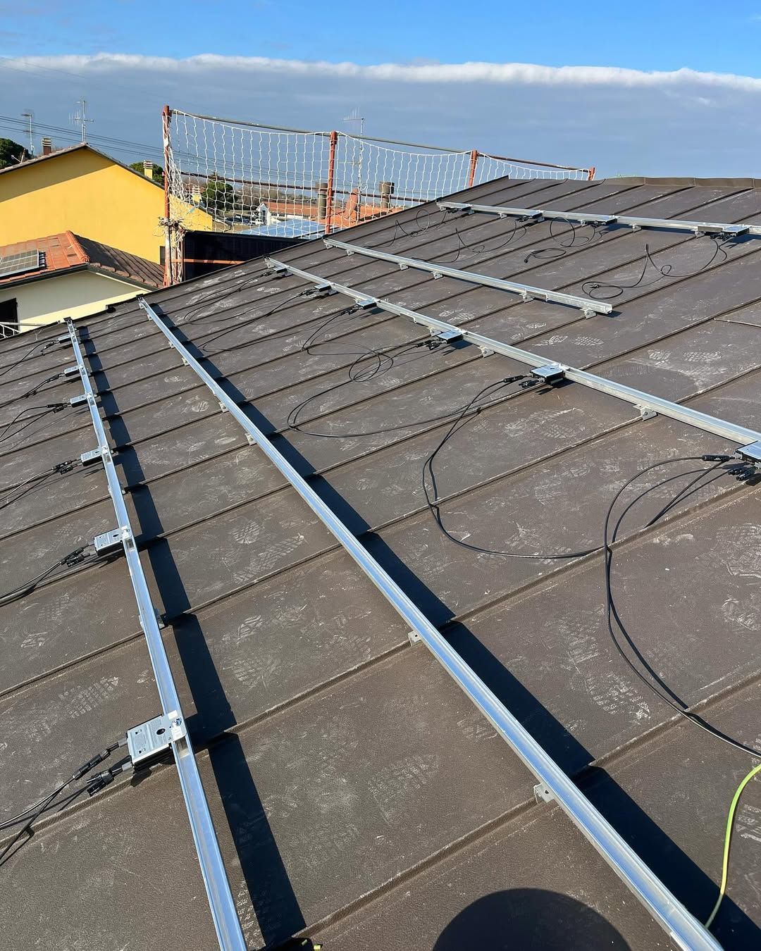 Binari di montaggio metallici per pannelli solari installati su un tetto di tegole scure, con i cavi elettrici a vista.