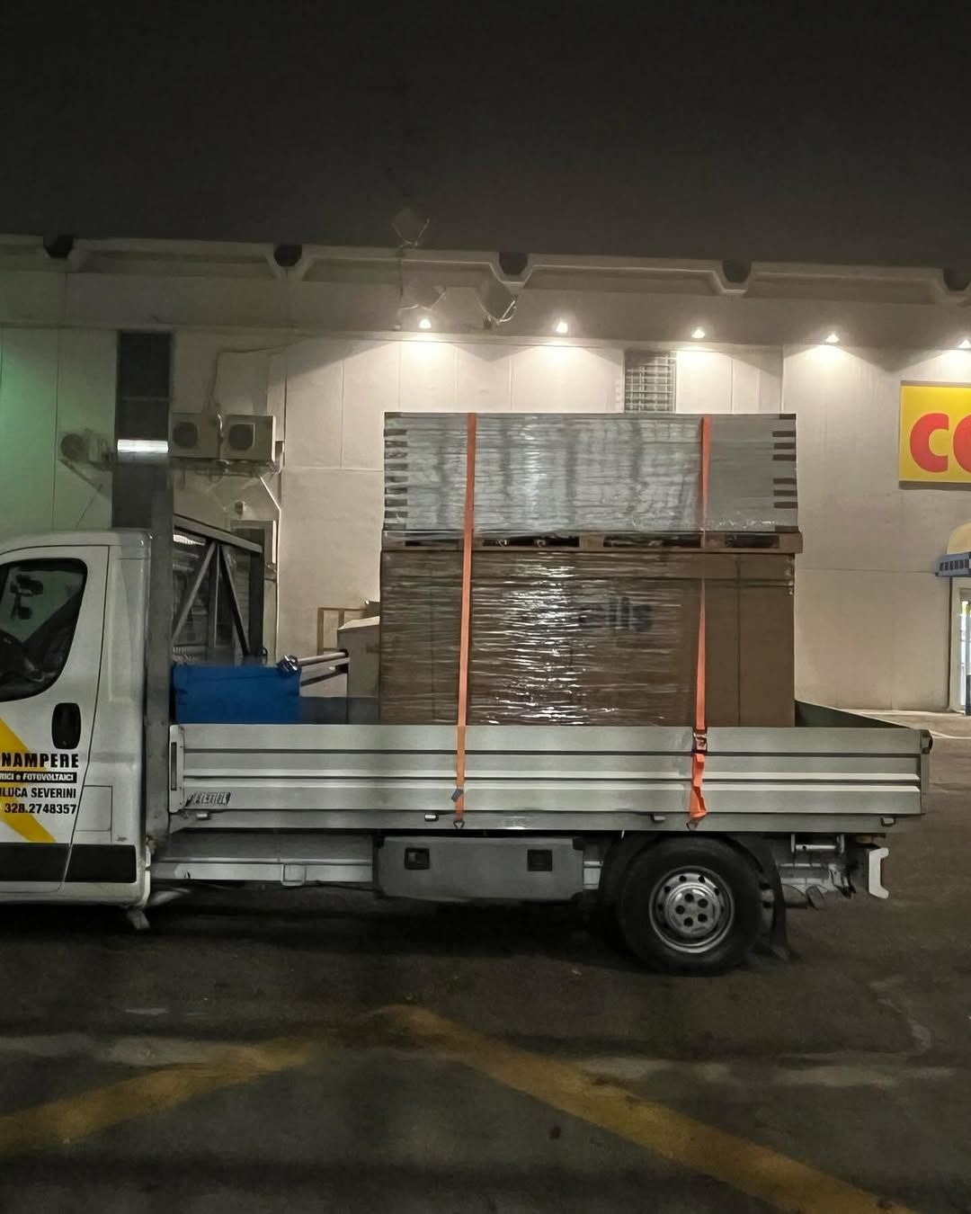 Un camioncino bianco a pianale ribassato parcheggiato di notte, che trasporta un grosso carico pallettizzato fissato con due cinghie di fissaggio arancioni.