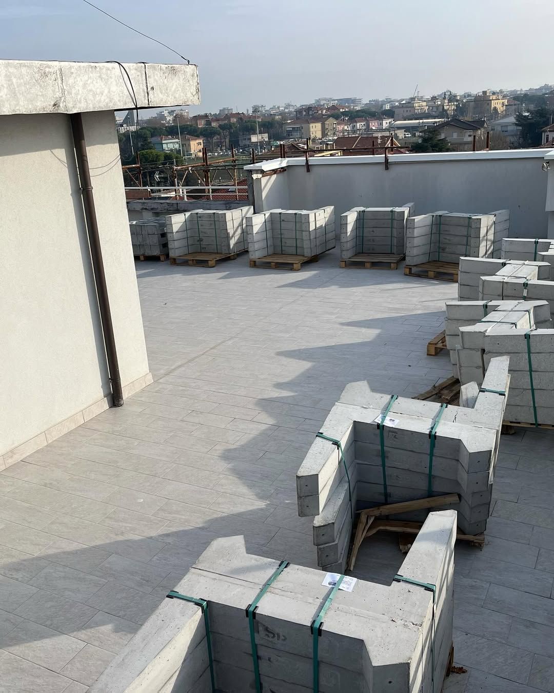 Pallet di blocchi di cemento grigio chiaro impilati e disposti su un tetto, con uno scorcio di città sullo sfondo.