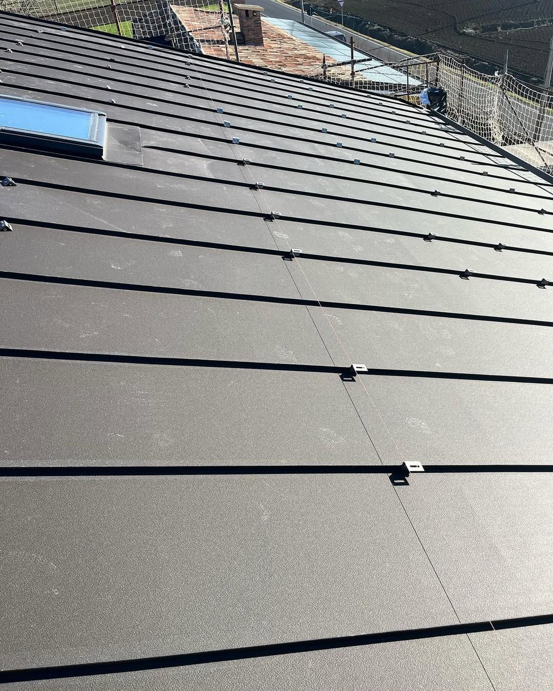 Primo piano di un tetto in metallo scuro con giunture orizzontali e piccole clip di fissaggio, che mostra un lucernario in alto a sinistra.