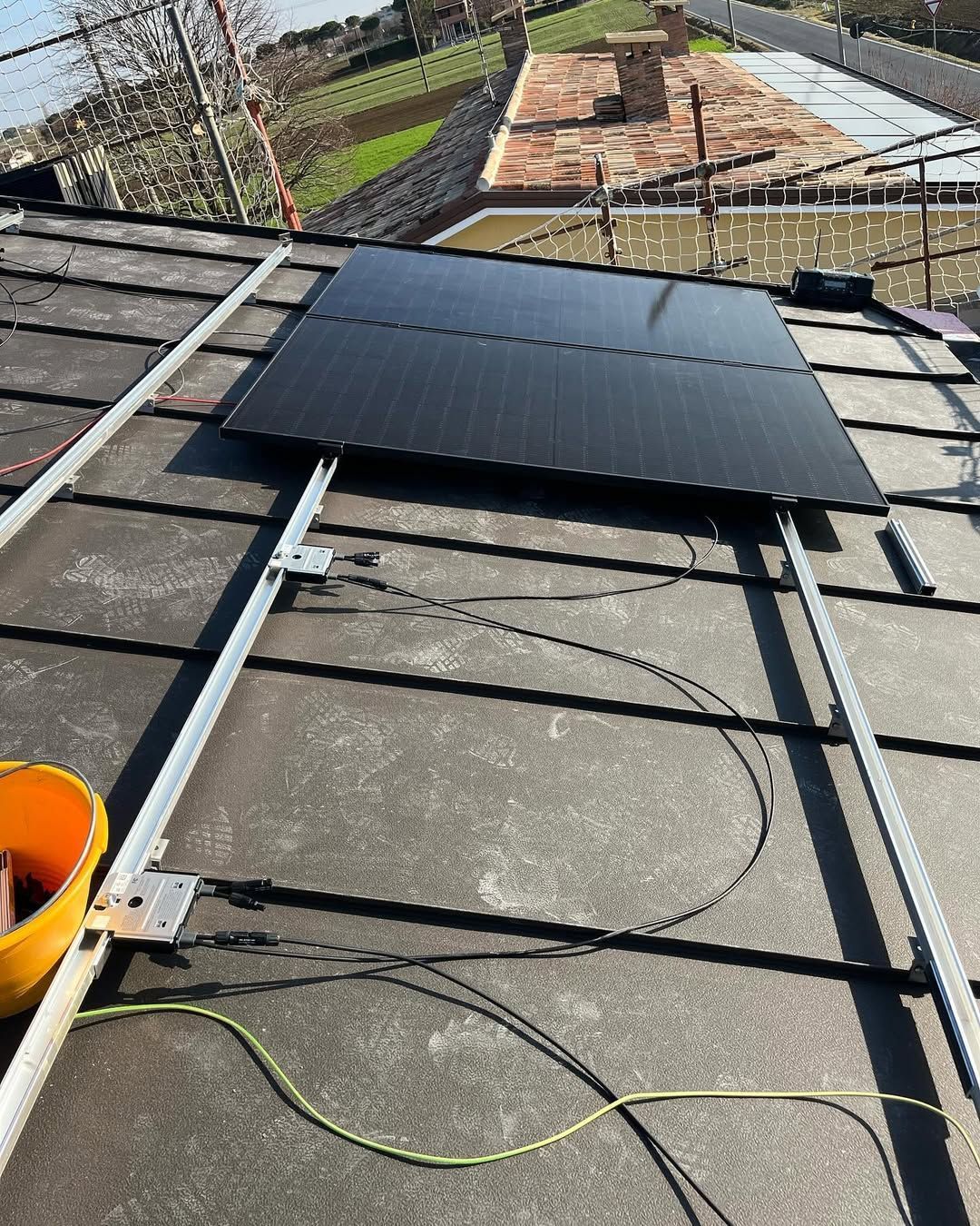 Un pannello solare nero montato su binari metallici fissati a un tetto di tegole scure, con cavi elettrici arrotolati sulla superficie.
