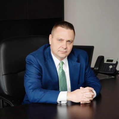 A man in a blue suit and green tie is sitting at a desk with his hands folded.