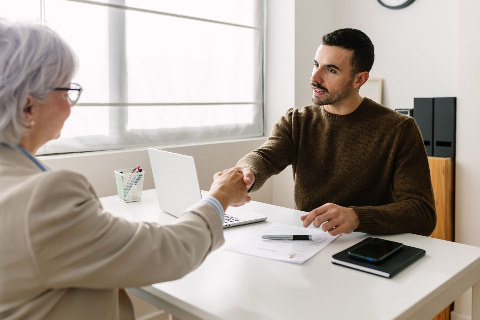 Two people shaking hands across a desk with a laptop and documents.