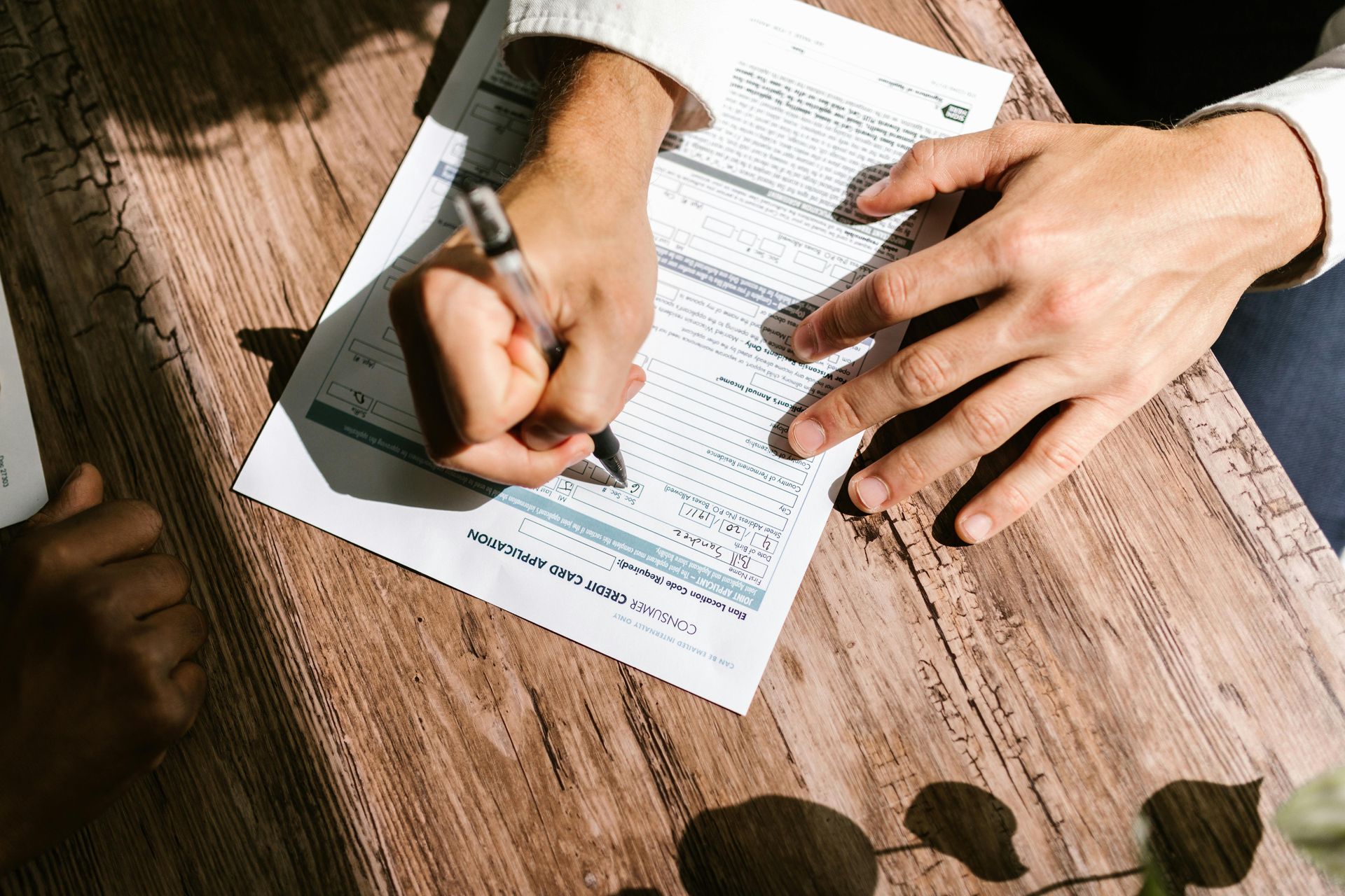 Person writing on a form with a pen, on a wooden table.