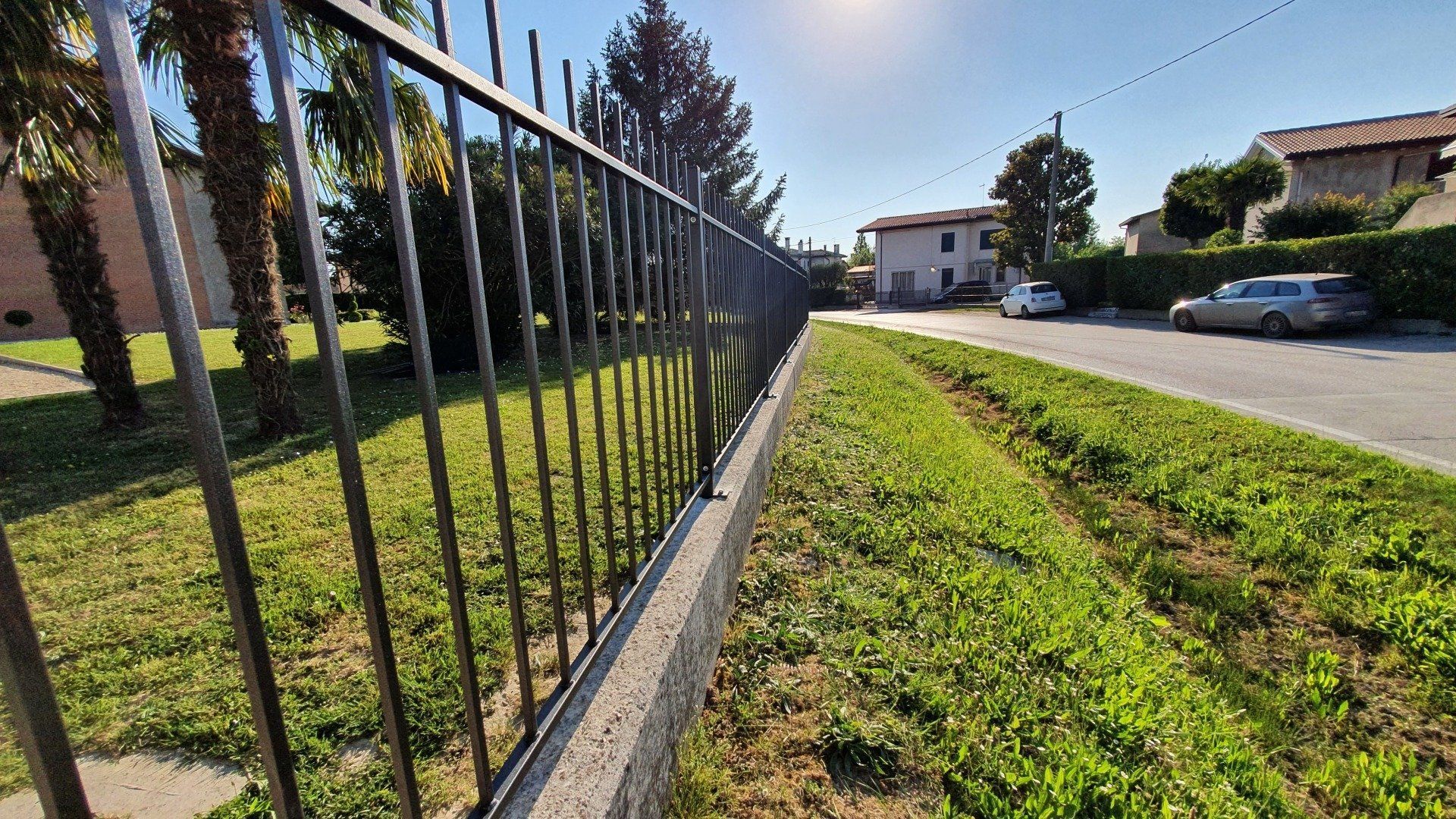 Recinzione metallica nera che delimita un'area erbosa accanto a una strada, con case e automobili visibili sullo sfondo.