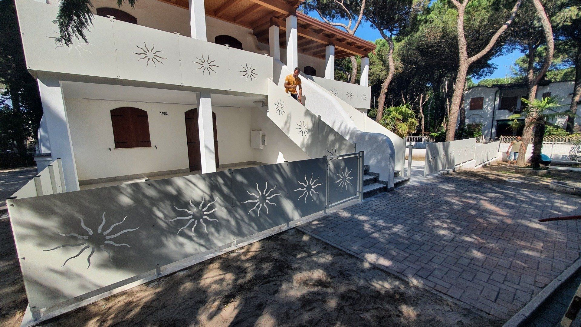 Edificio bianco con ringhiera a raggiera, gradini e persiane marroni. Posizione esterna soleggiata.