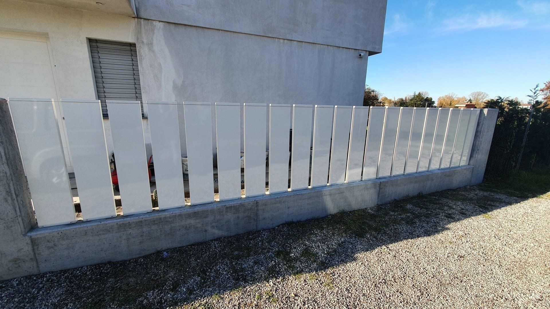 Staccionata bianca di fronte a un muro e a un edificio in cemento, contro un cielo azzurro.