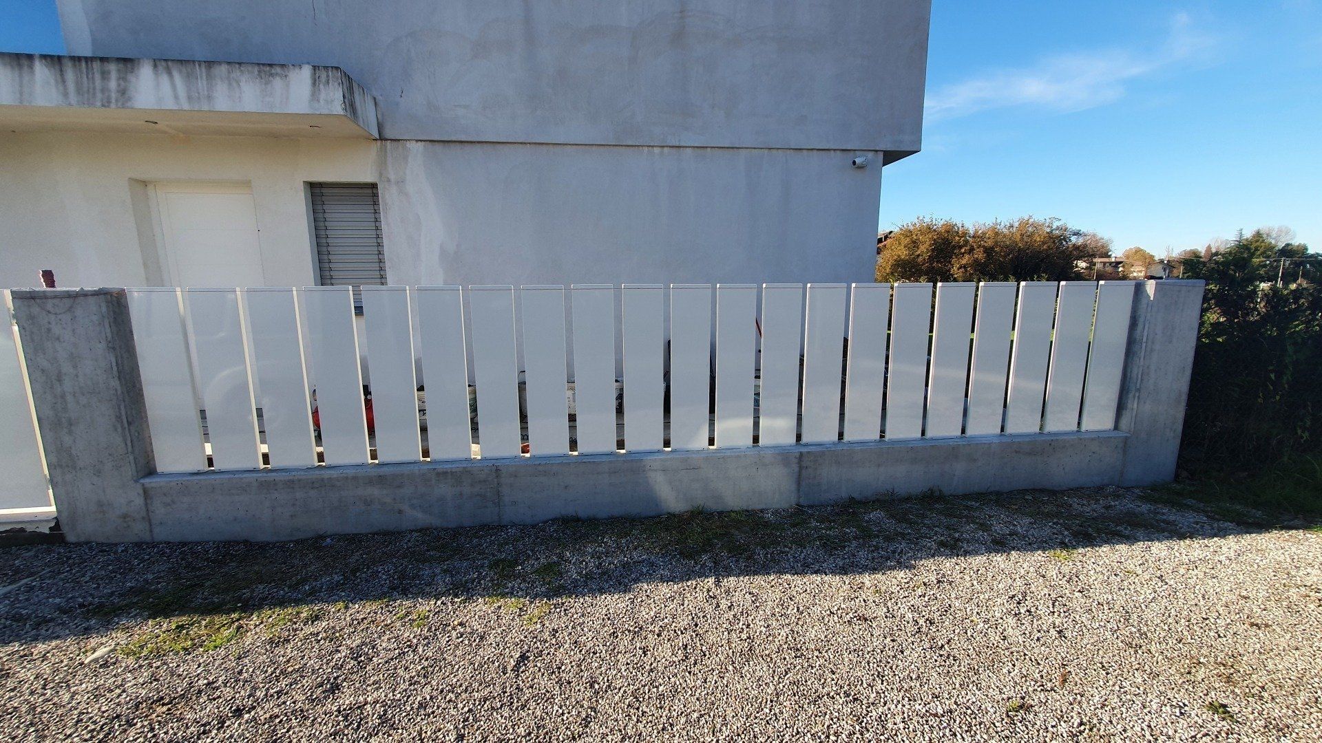 Staccionata bianca con pilastri di cemento di fronte a un edificio bianco, su ghiaia.
