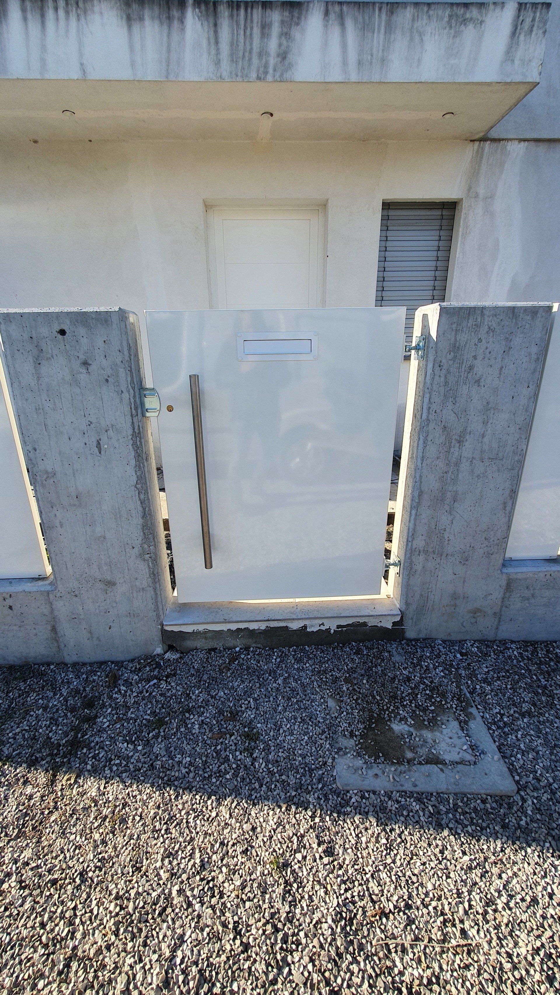 Cancello di metallo bianco tra due pilastri di cemento, di fronte a un edificio con una porta bianca.