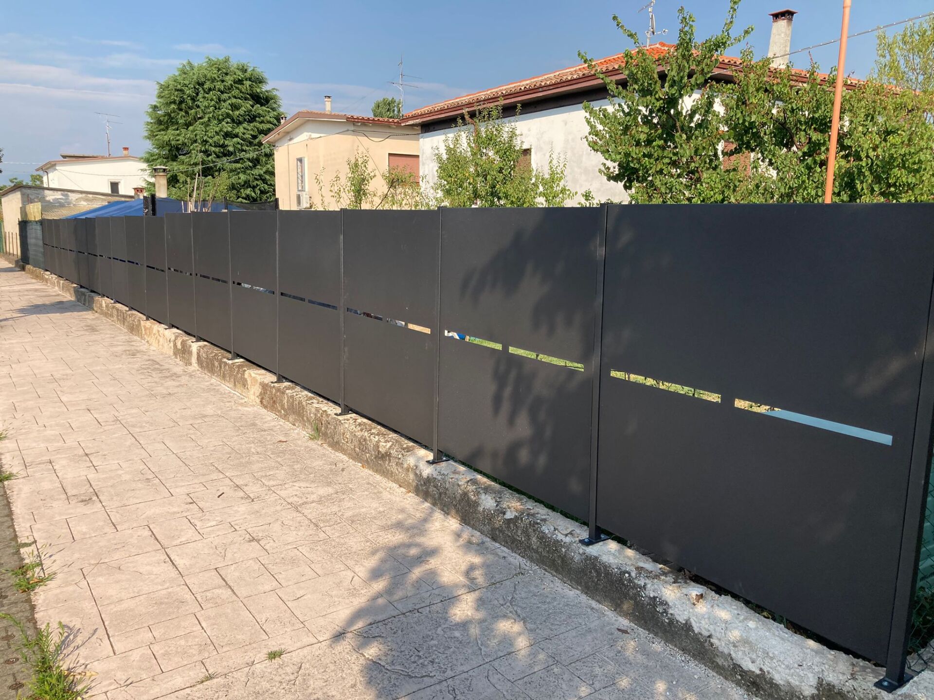 Moderna recinzione grigio scuro lungo un marciapiede in mattoni, con edifici e alberi sullo sfondo.