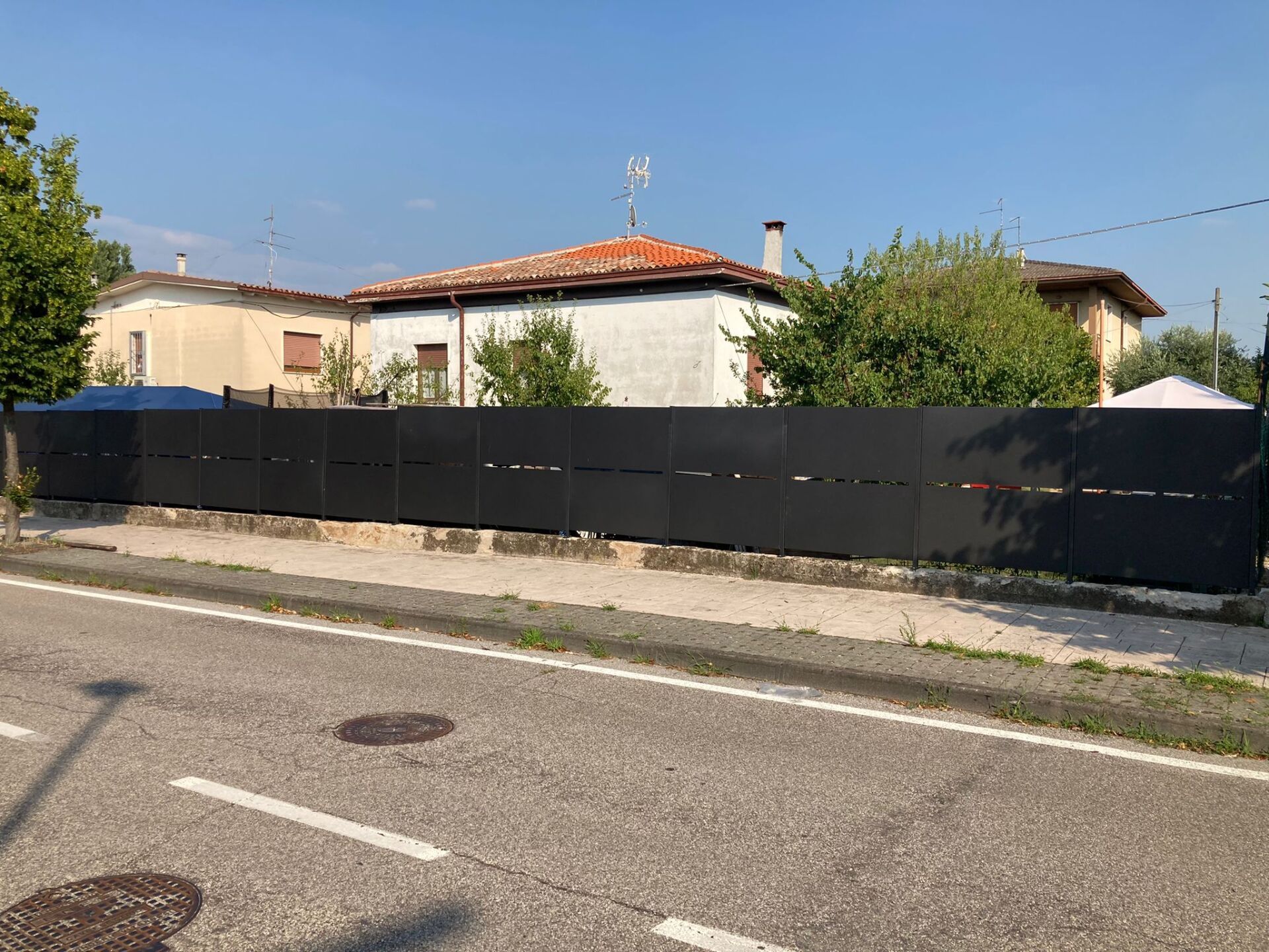 Recinzione nera lungo una strada, di fronte a una fila di edifici e alberi sotto un cielo limpido.