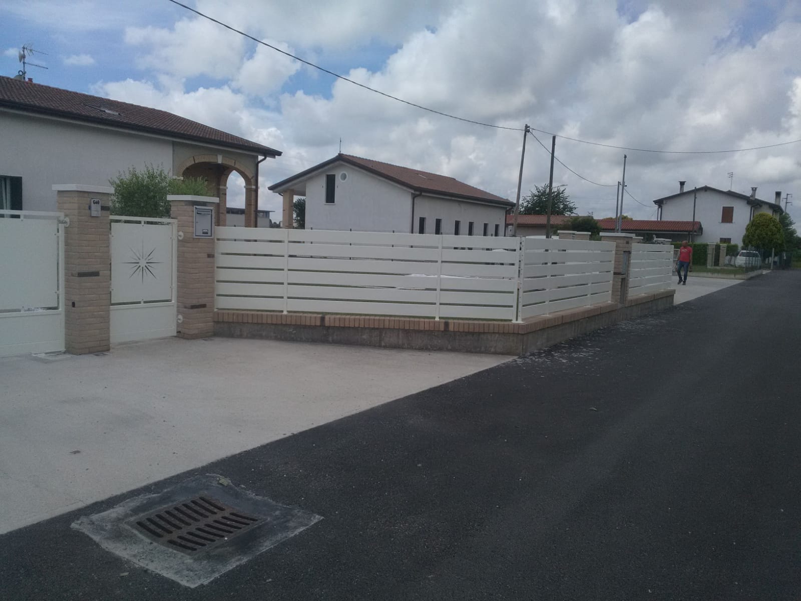 Recinzione bianca con dettagli in mattoni lungo una strada asfaltata, case sullo sfondo. Cielo coperto.