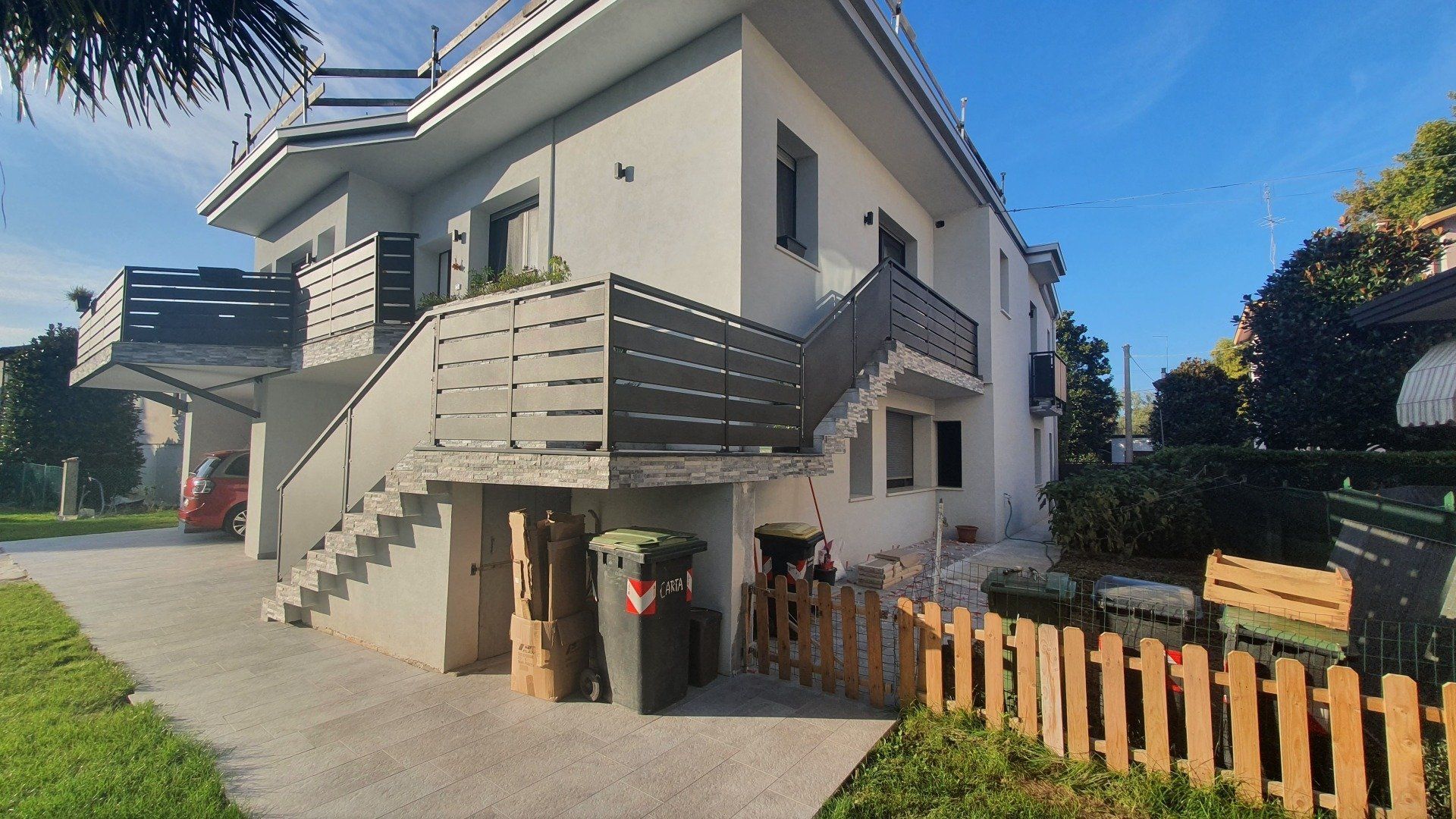 Esterno di un edificio a più piani con balconi, scale e una piccola recinzione in legno; pareti grigio chiaro.