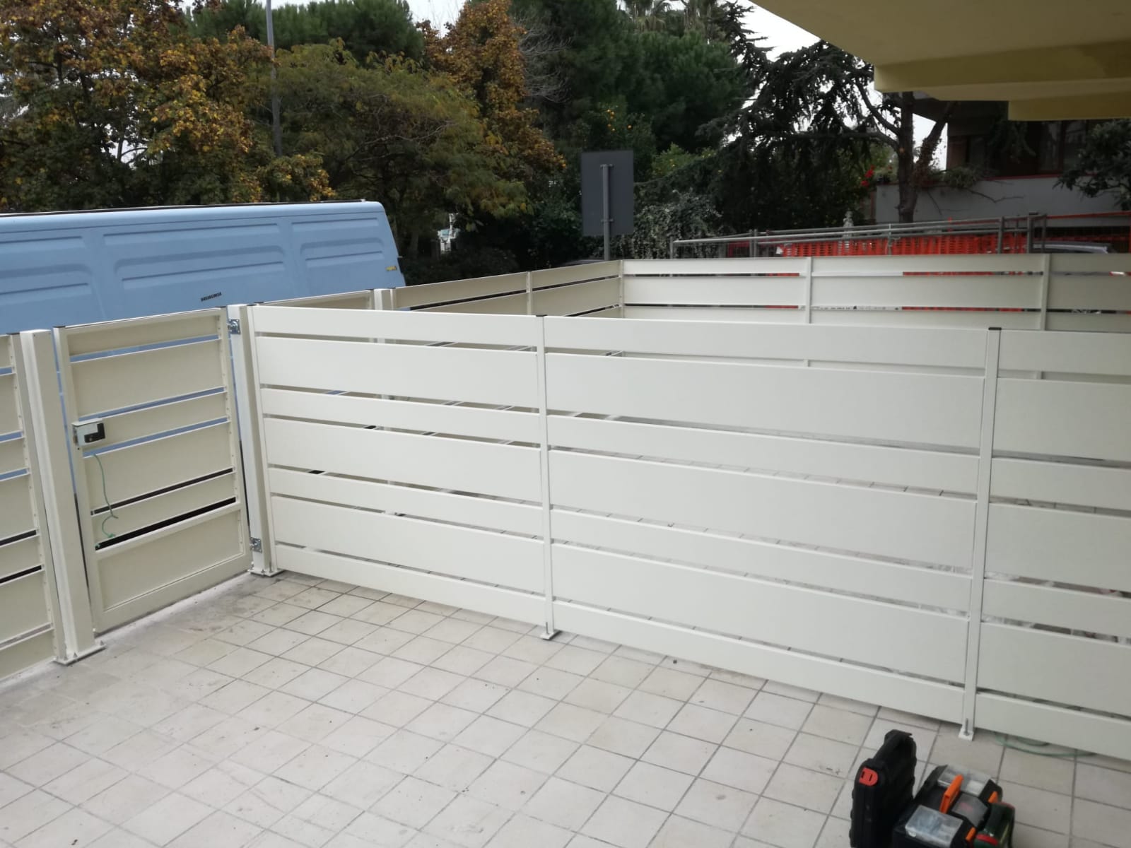 Recinzione e cancello in listelli bianchi su un patio piastrellato. Sullo sfondo si vedono alberi e un veicolo.