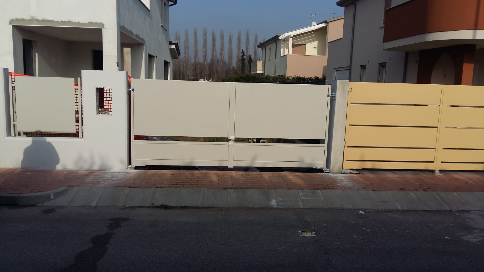 Cancello e recinzione in metallo color marrone chiaro di fronte a case chiare e a una fila di alberi.