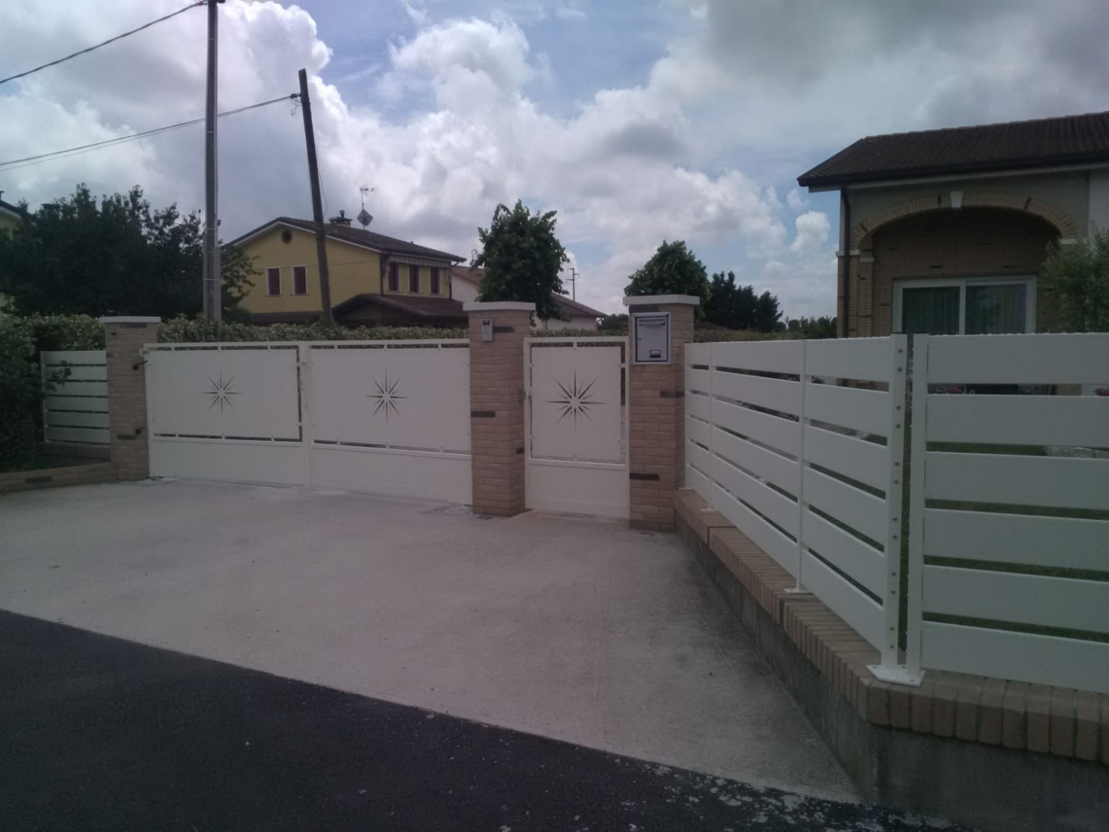 Un cancello bianco e una recinzione delimitano un vialetto. Sullo sfondo, pilastri in mattoni e una casa. Cielo nuvoloso.