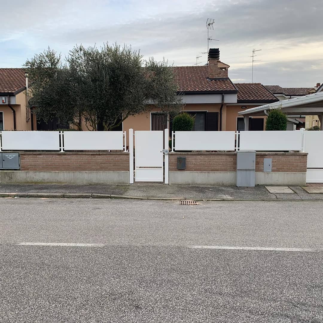 Recinzione bianca e cancello davanti a una casa in mattoni e stucco, con un albero e un cielo coperto.