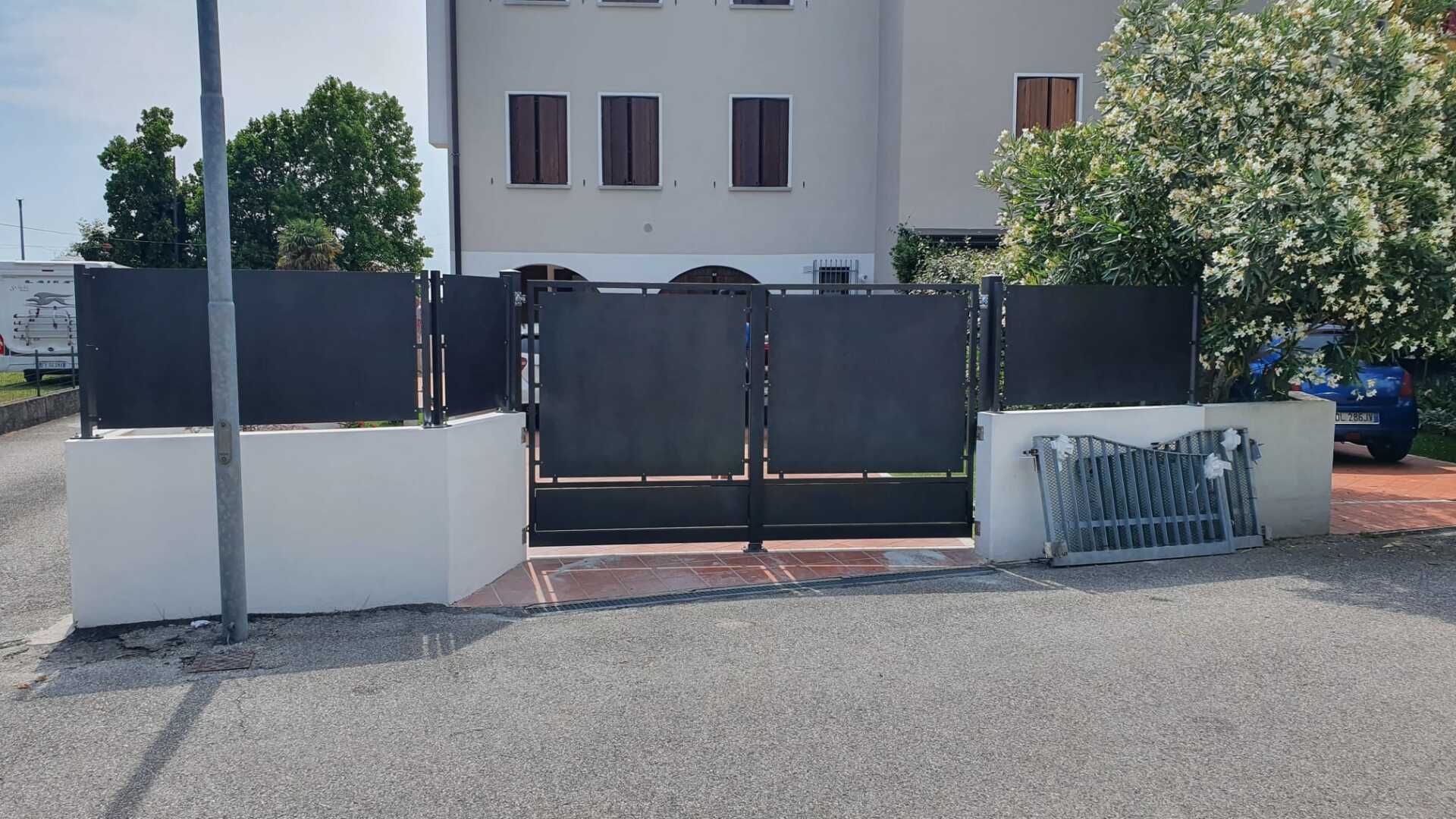Cancello e recinzione in metallo nero di fronte a un edificio bianco con alberi e cielo azzurro.