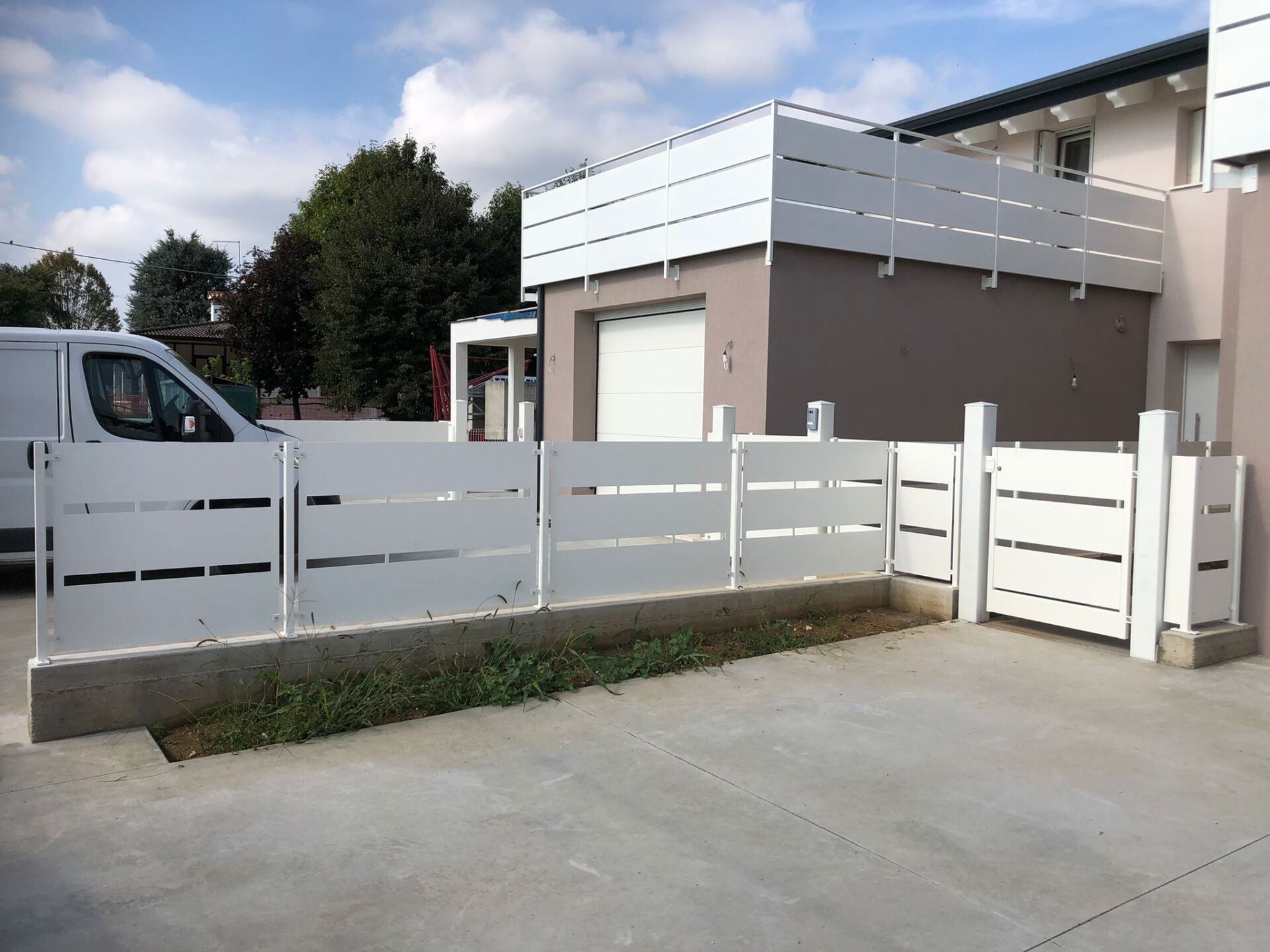 Recinzione bianca con stecche orizzontali davanti a una casa moderna, vialetto in cemento, cielo coperto.