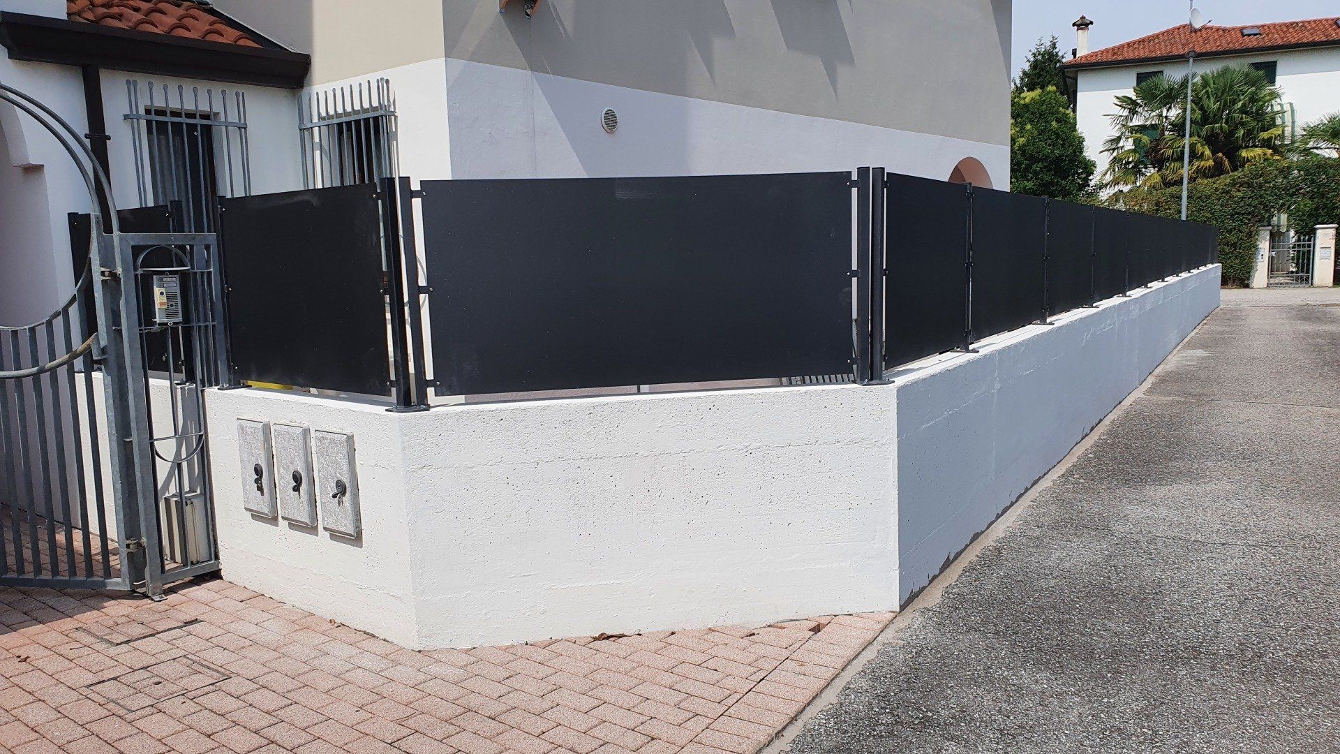 Recinzione scura in cima a un muro di cemento lungo l'angolo di un edificio, su un sentiero di mattoni e cemento.