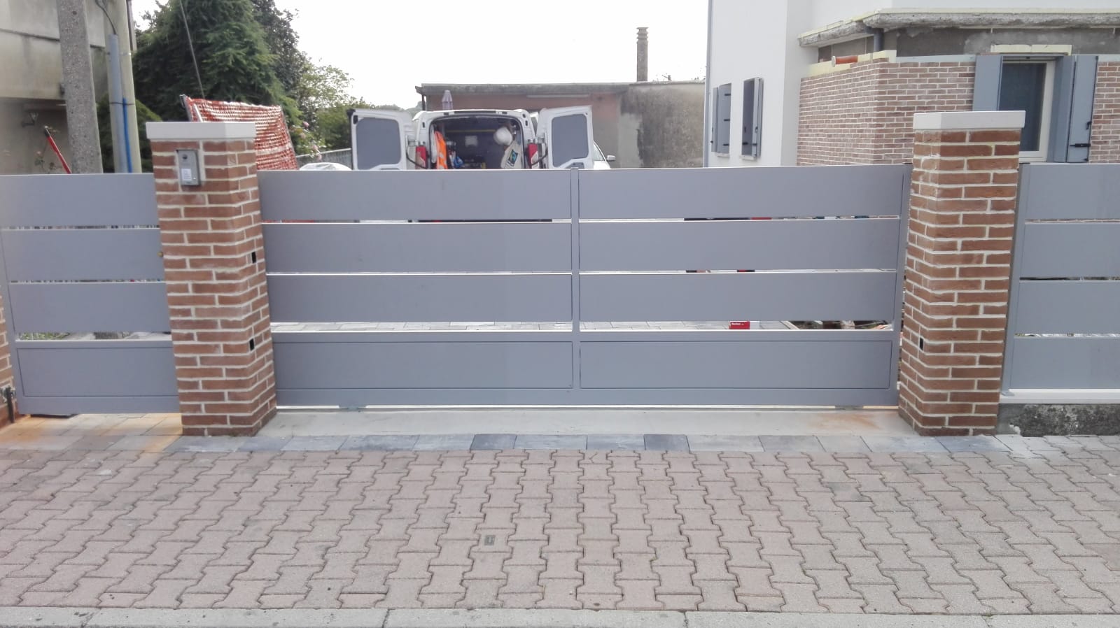 Cancello in metallo grigio con pilastri in mattoni. Il cancello si chiude su un vialetto lastricato in mattoni.