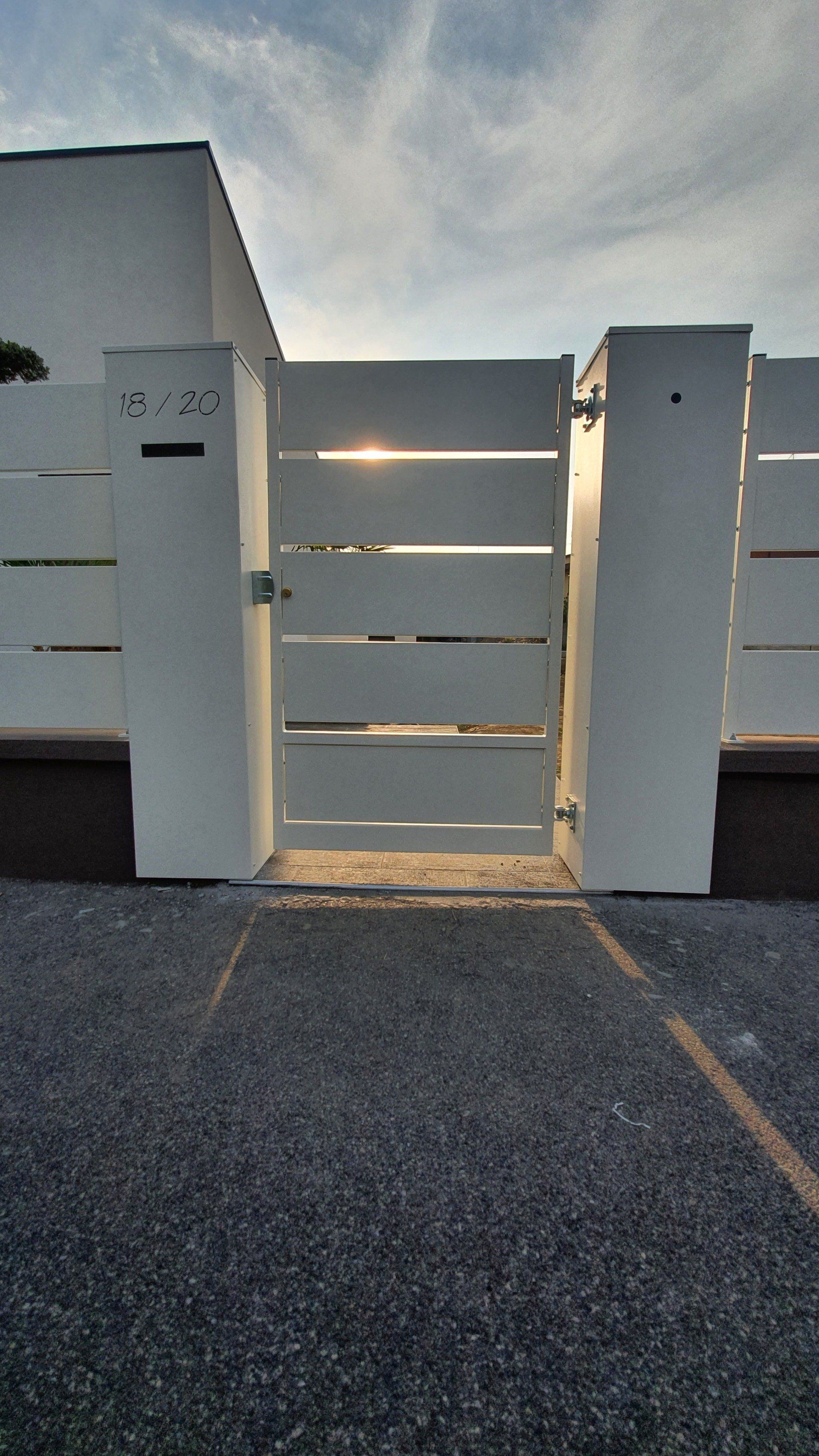 Cancello bianco con stecche orizzontali, tra due pilastri, di fronte a un edificio bianco, su un sentiero di ghiaia.