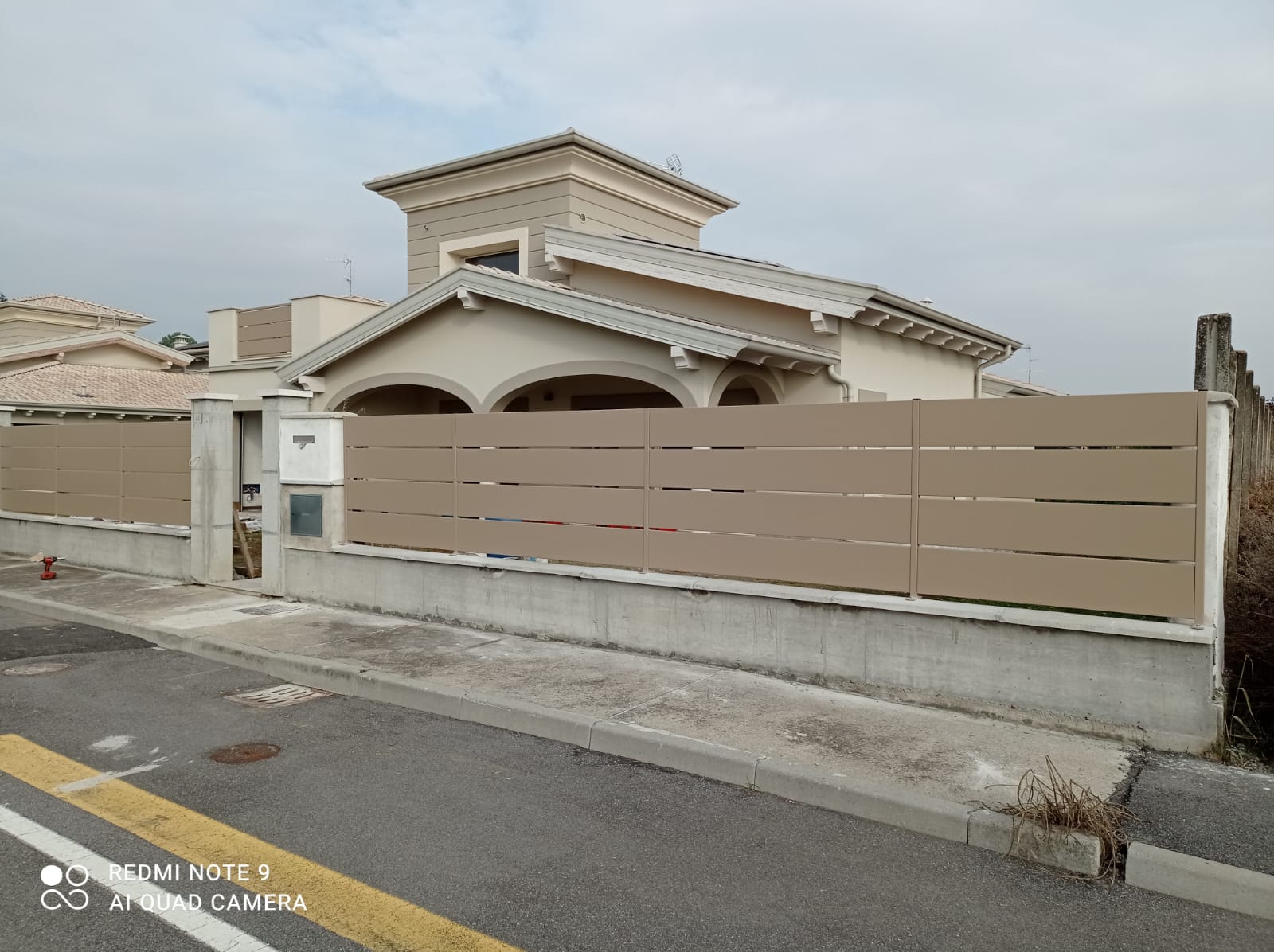 Recinzione in doghe color marrone chiaro su una base di cemento di fronte a una casa beige con tetto di tegole.