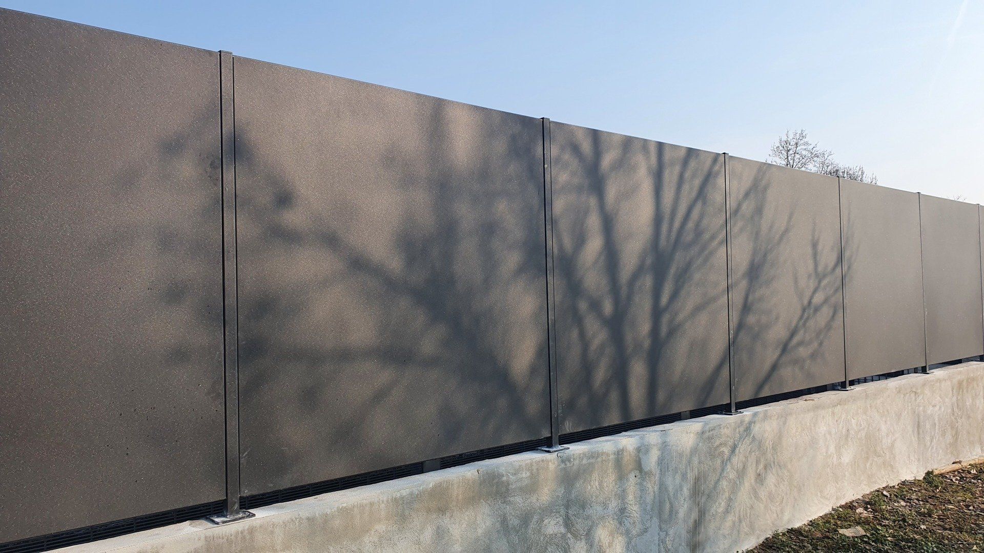 Recinzione dalla trama scura contro un cielo azzurro, che proietta le ombre degli alberi sui pannelli.