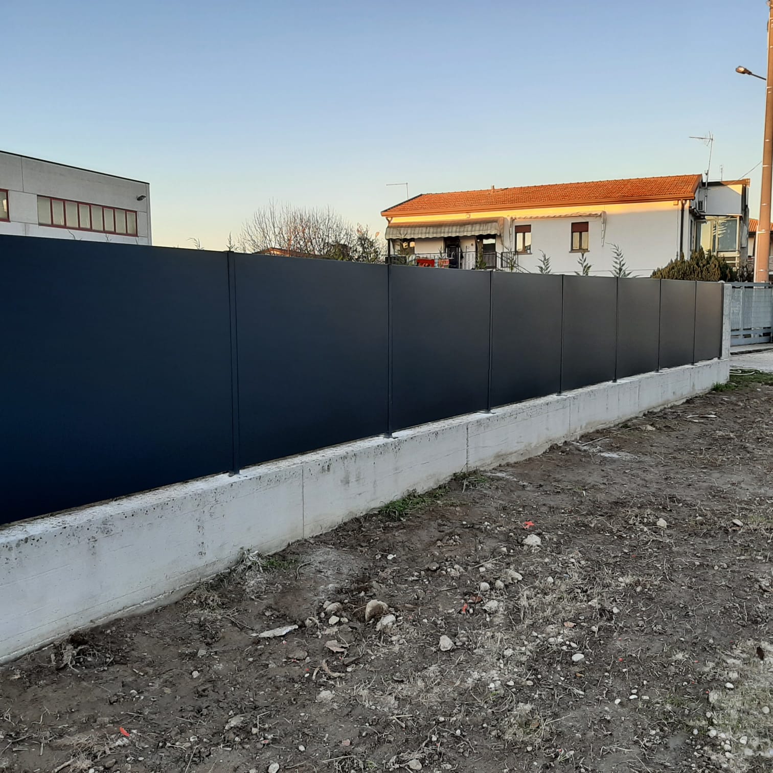 Recinzione grigio scuro su una base di cemento, che separa un terreno sterrato dagli edifici sullo sfondo.