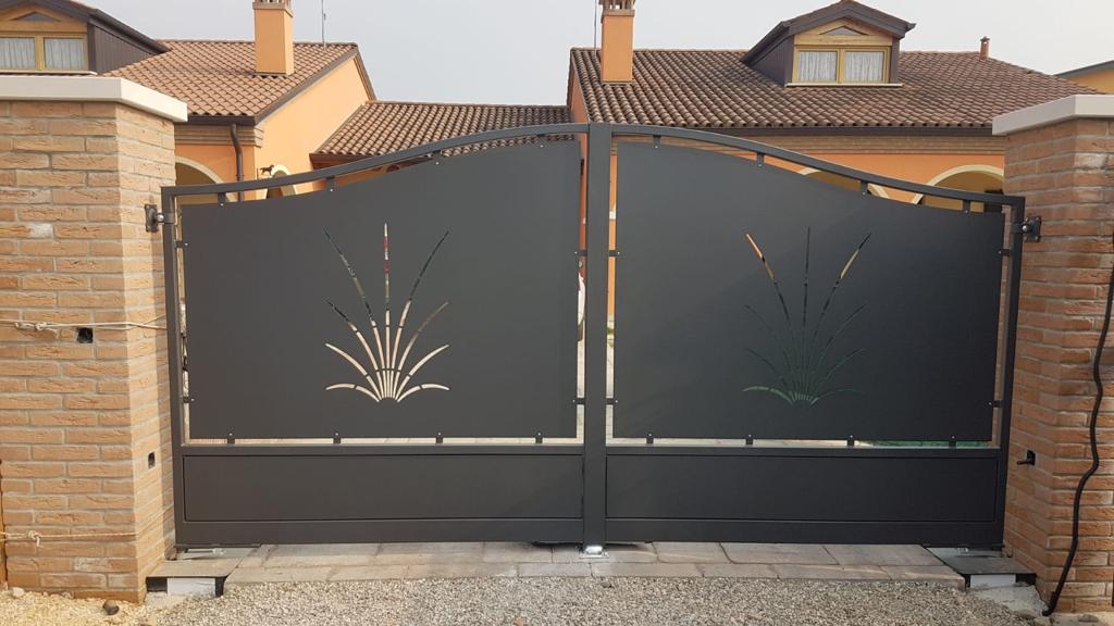 Cancello d'accesso in metallo scuro con ritagli decorativi, inserito tra pilastri in mattoni di fronte a una casa.