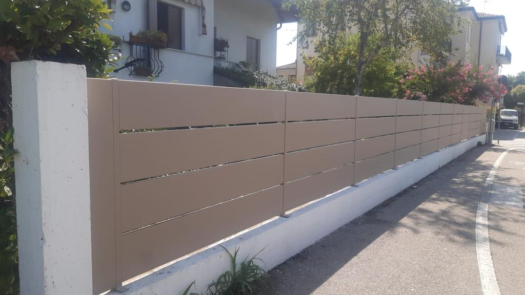 Recinzione beige con listelli orizzontali in cima a un muro bianco lungo una strada.