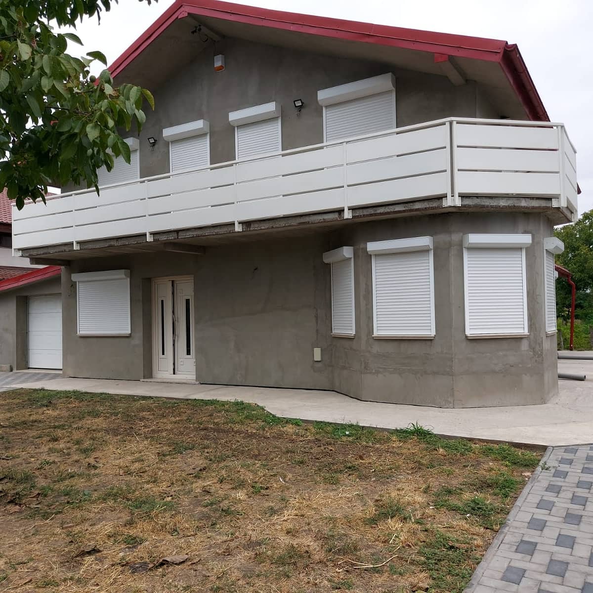 Casa in stucco grigio con tetto rosso, persiane bianche, balcone e porta d'ingresso.
