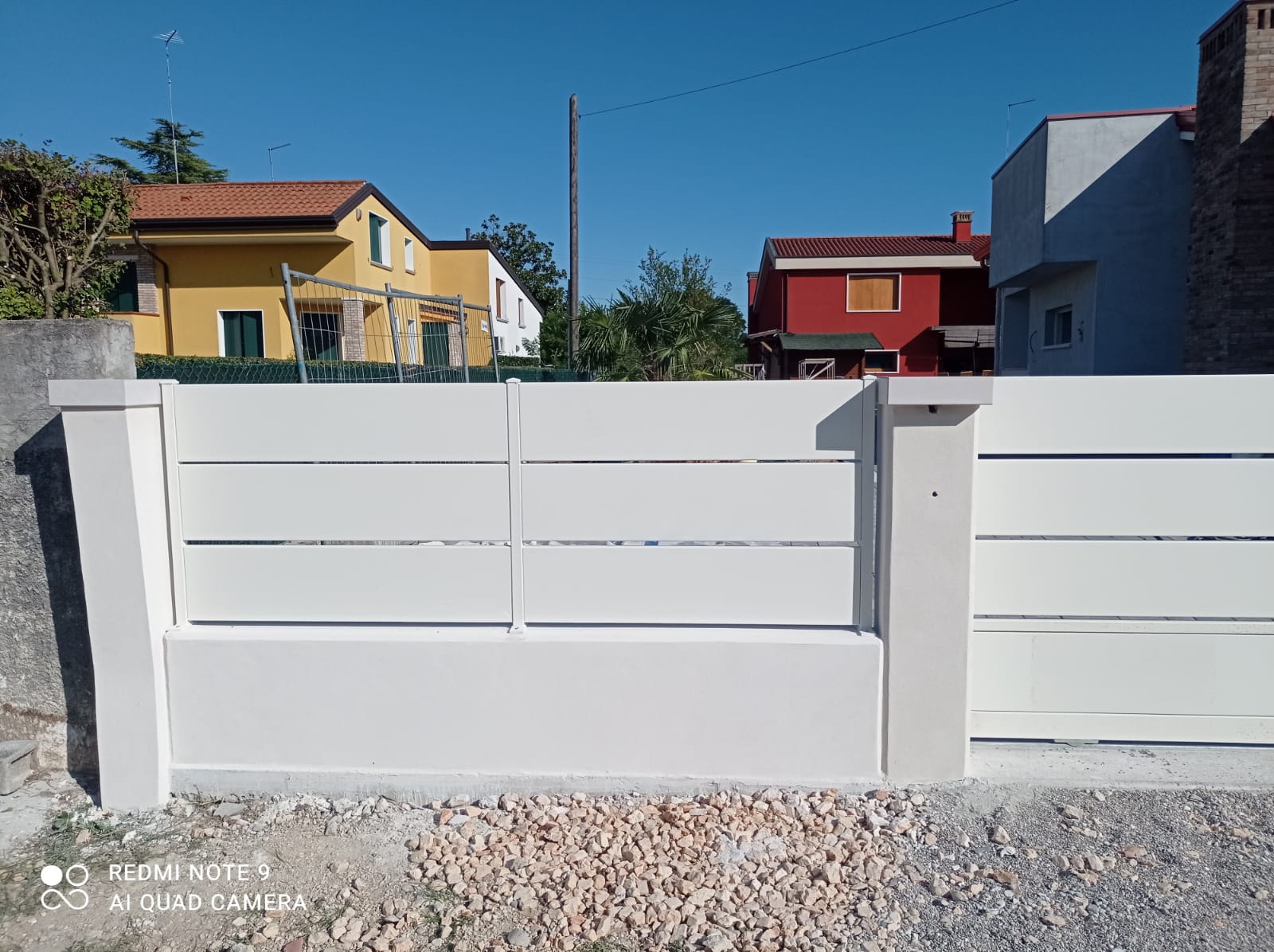 Recinzione bianca a doghe orizzontali con due case sullo sfondo. Ghiaia e cielo azzurro.
