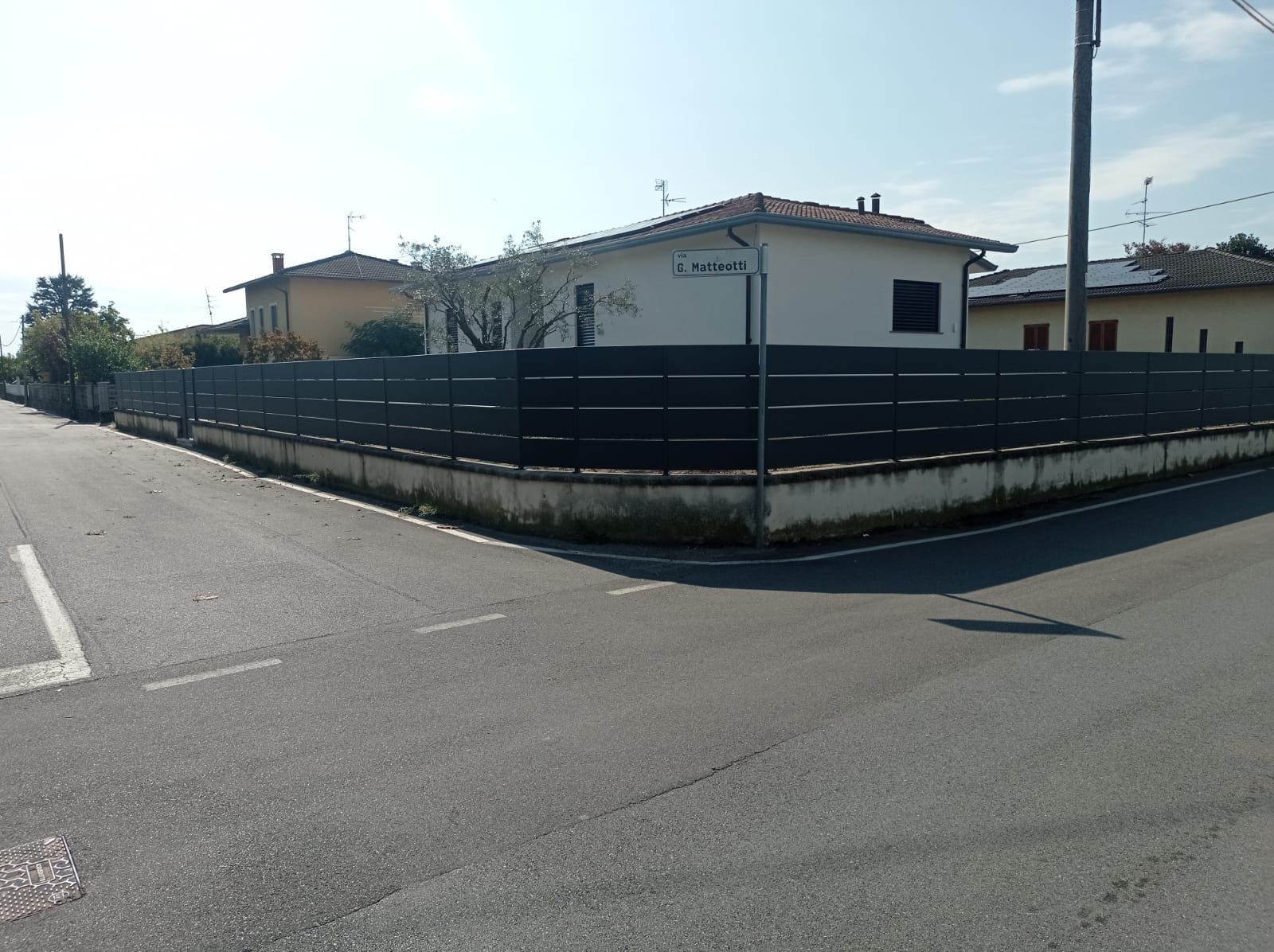 Angolo di strada con una recinzione grigio scuro, una casa bianca e un cartello stradale sotto un cielo limpido.