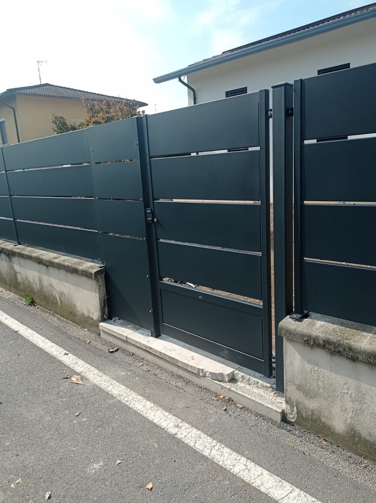 Recinzione in listelli orizzontali grigio scuro con cancello, base in cemento e marciapiede.