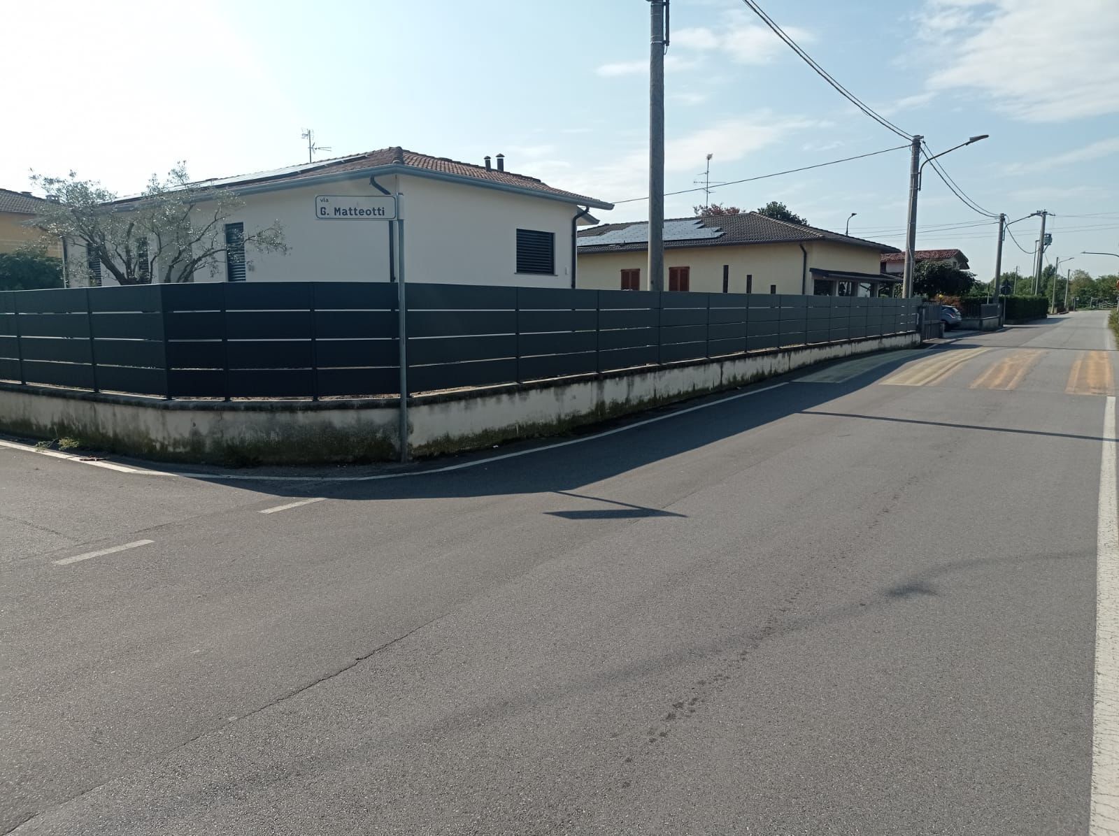 Una strada asfaltata curva dietro un angolo con case e una recinzione scura. In alto, linee elettriche e un cielo limpido.