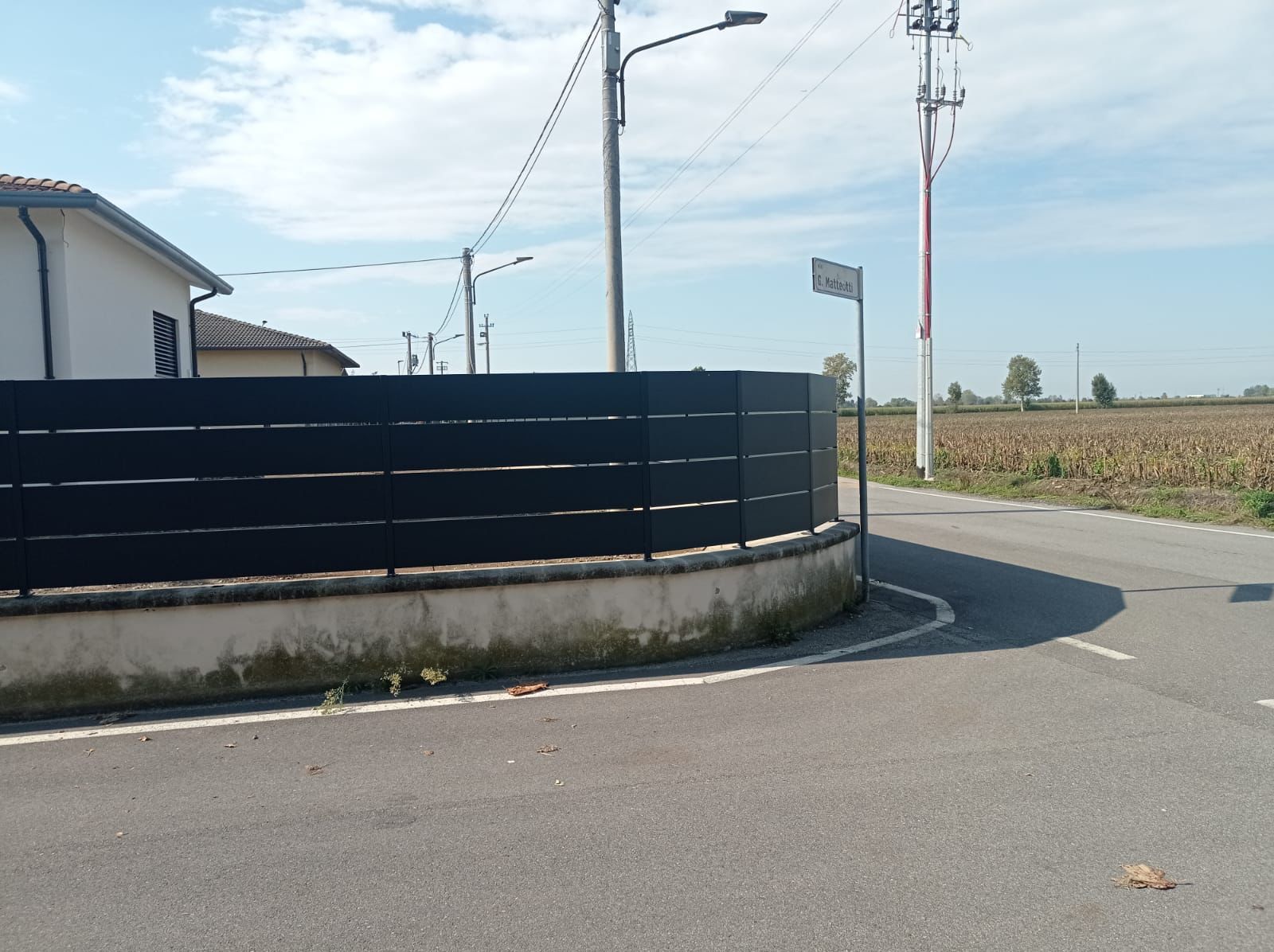 Un angolo di strada con una strada curva, una recinzione scura, un cartello e pali della luce sotto un cielo azzurro.