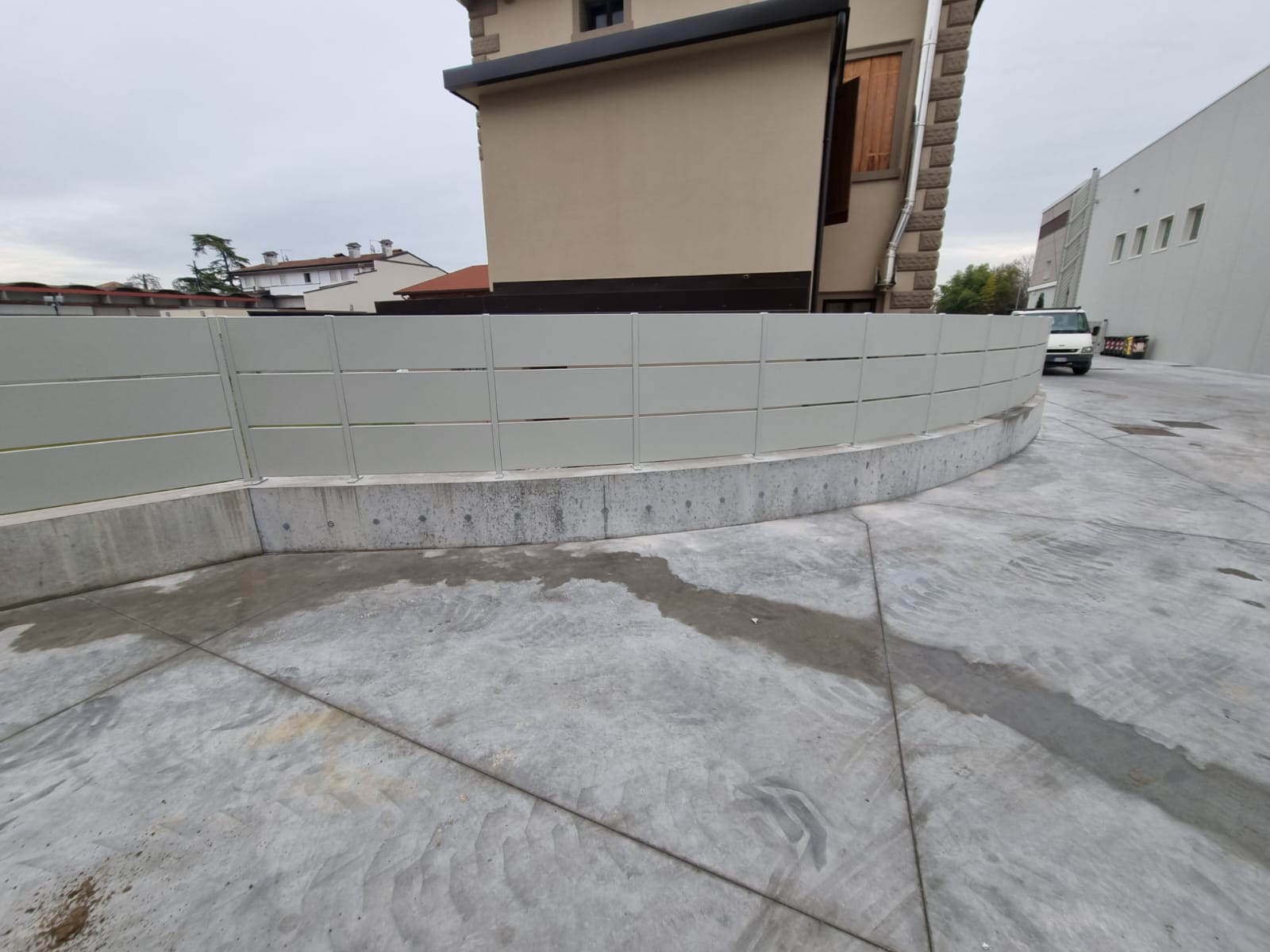 Un muro curvo di cemento con una recinzione grigia di fronte a un edificio beige in una giornata nuvolosa.