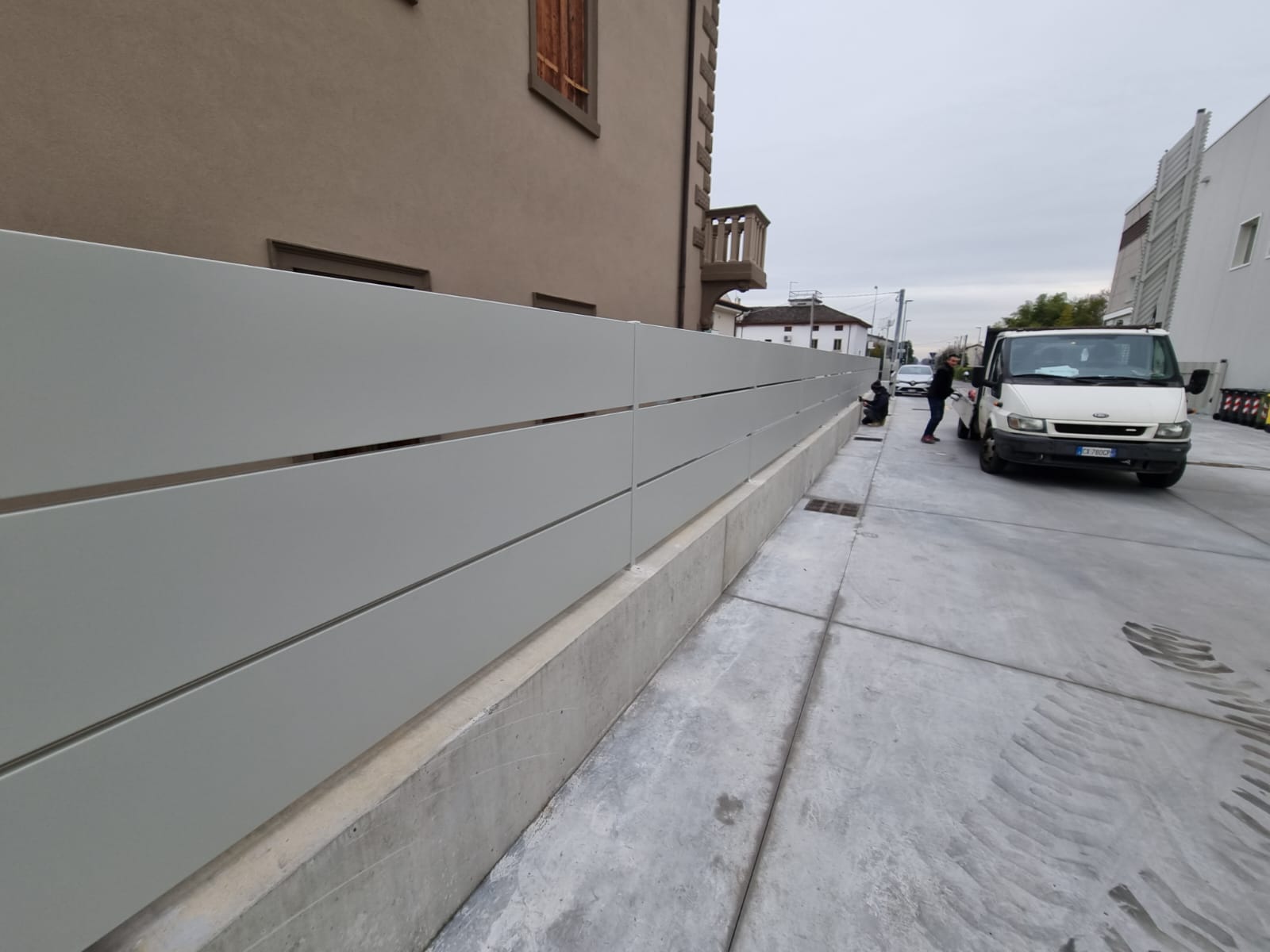 Recinzione orizzontale grigia lungo un marciapiede in cemento con un furgone parcheggiato nelle vicinanze.