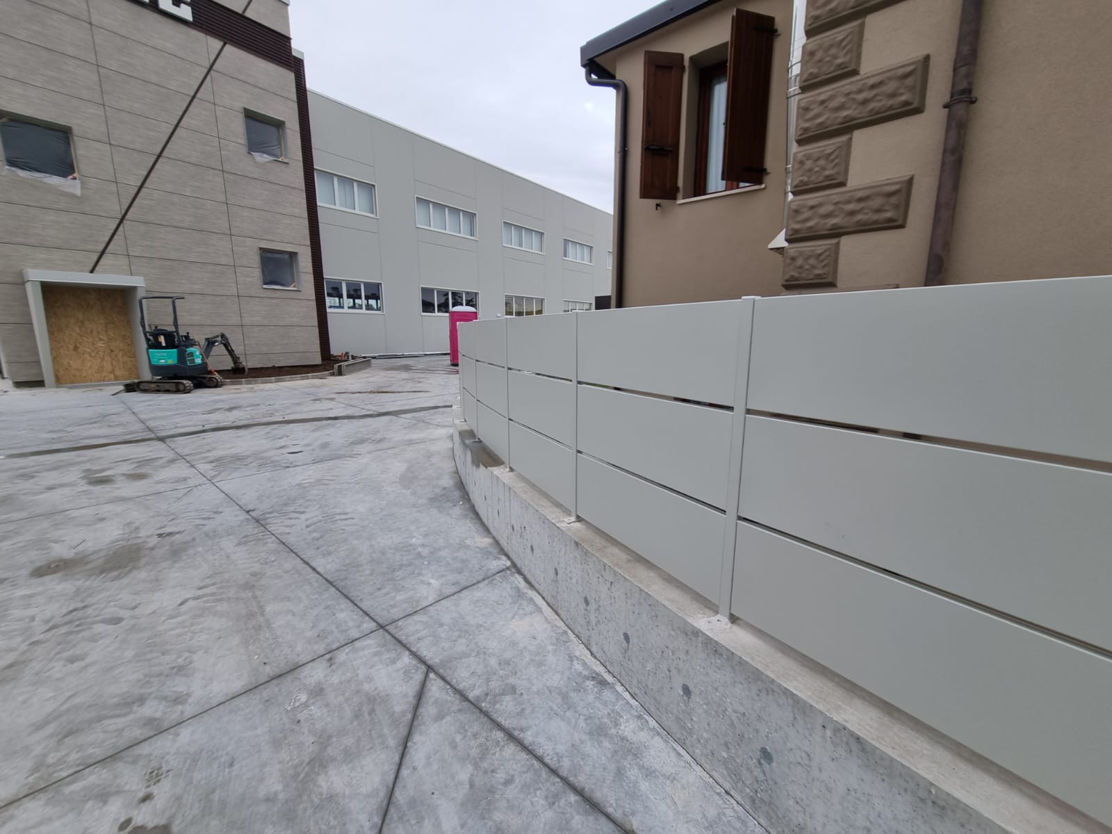 Recinzione metallica grigia curva, terreno in cemento, due edifici, giornata nuvolosa.