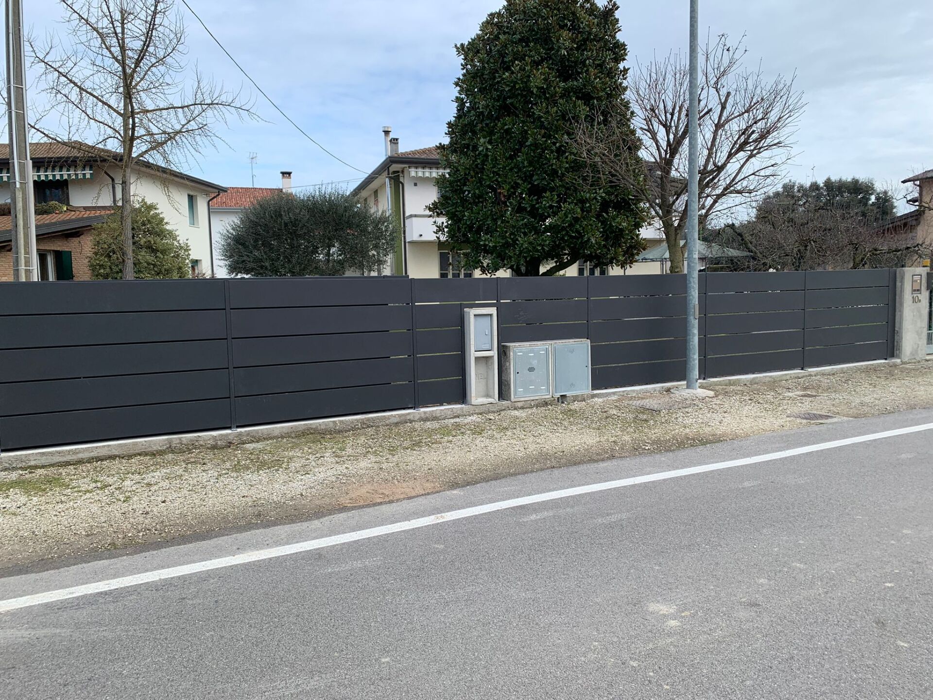 Una recinzione orizzontale grigio scuro lungo una strada, case sullo sfondo, cassette di servizio di fronte.