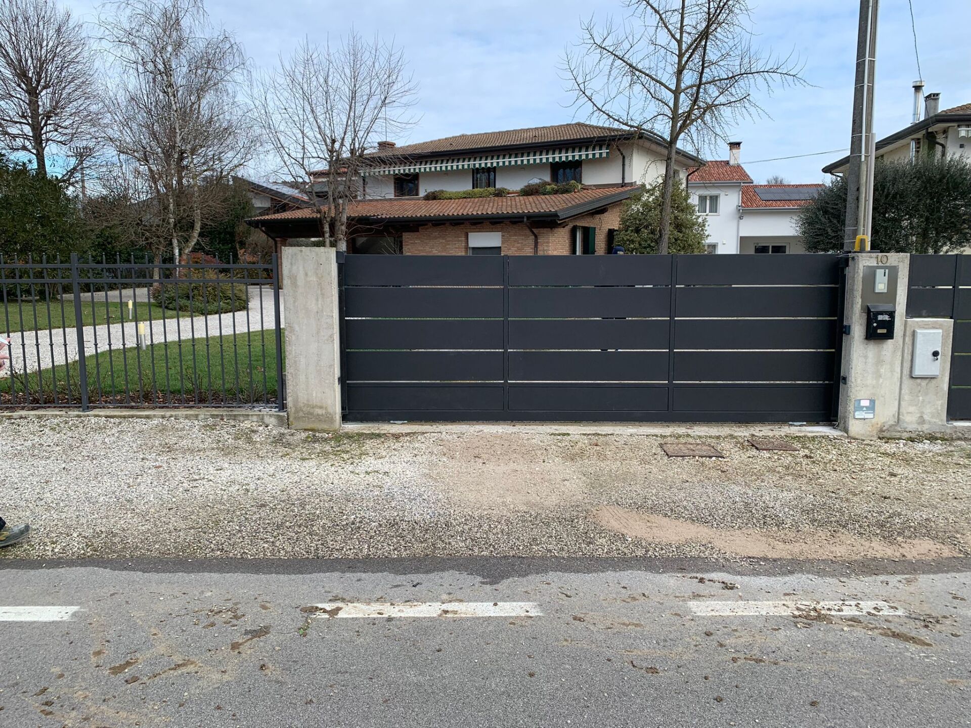 Cancello orizzontale grigio scuro davanti a una casa. Sono visibili pilastri di cemento e una piccola recinzione.