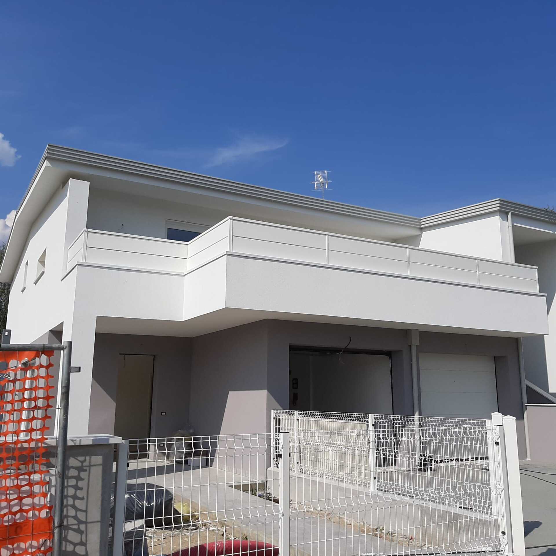 Casa bianca a due piani con balcone, pareti grigie, cielo azzurro. Recinzione di cantiere davanti.