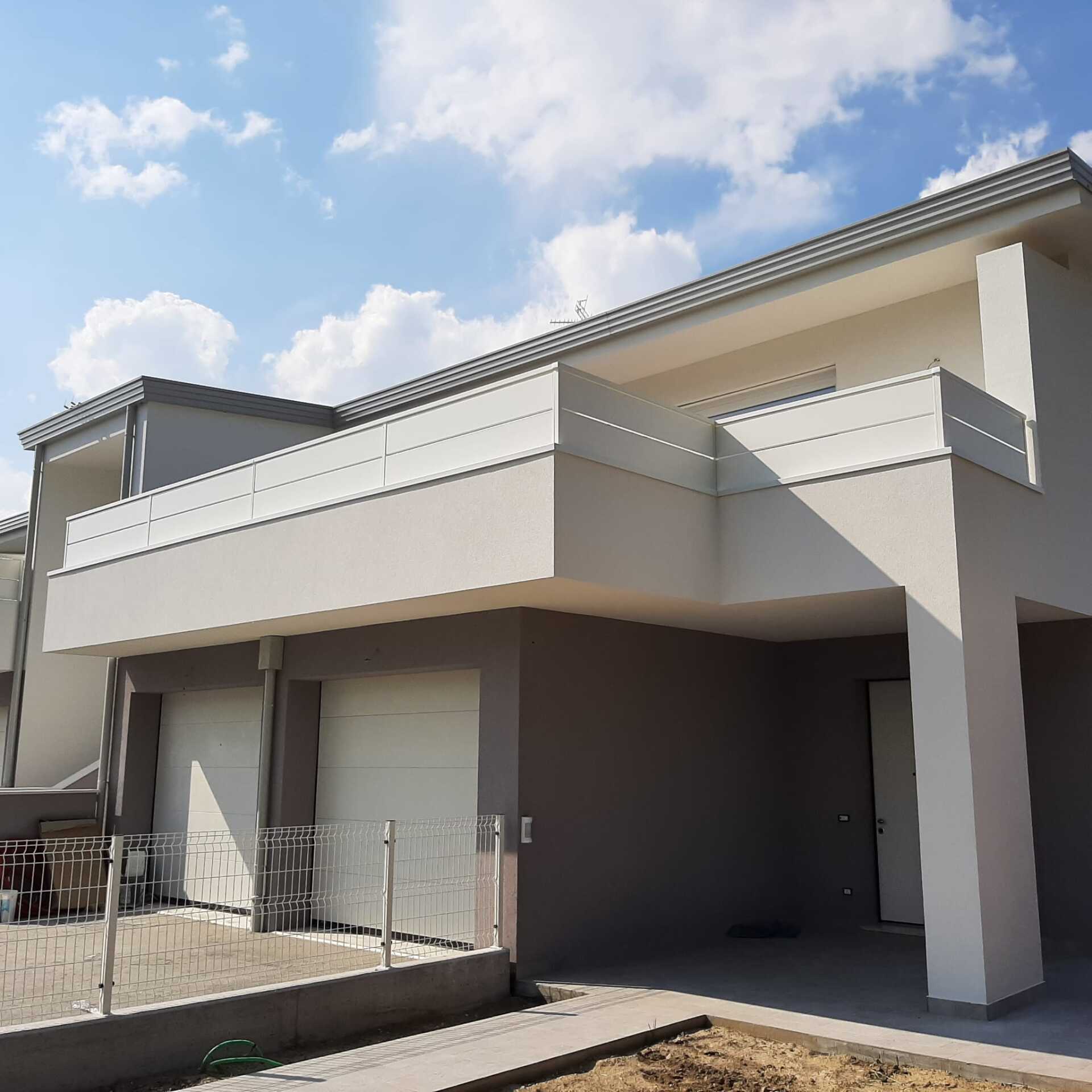 Moderna casa a due piani con balcone e porte da garage bianchi, esterni grigi e cielo azzurro.